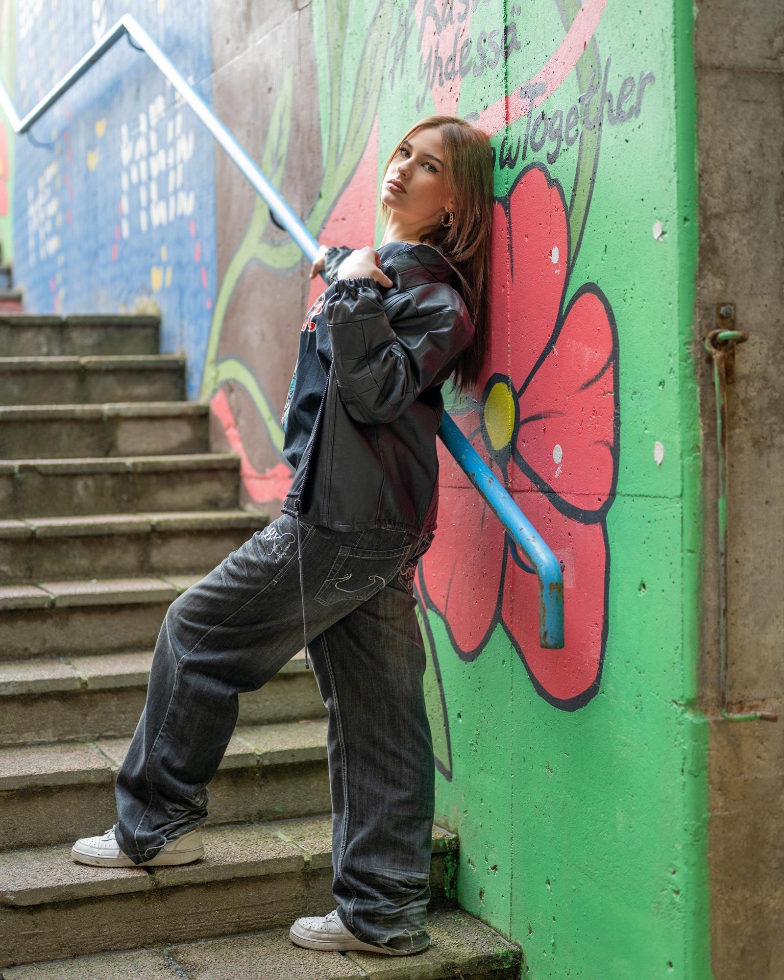 Young woman with long dark hair leans against a blue railing on concrete stairs, wearing a black jacket and dark jeans. Background features a colorful mural with a large red flower on a green and blue wall. White sneakers complete her outfit.