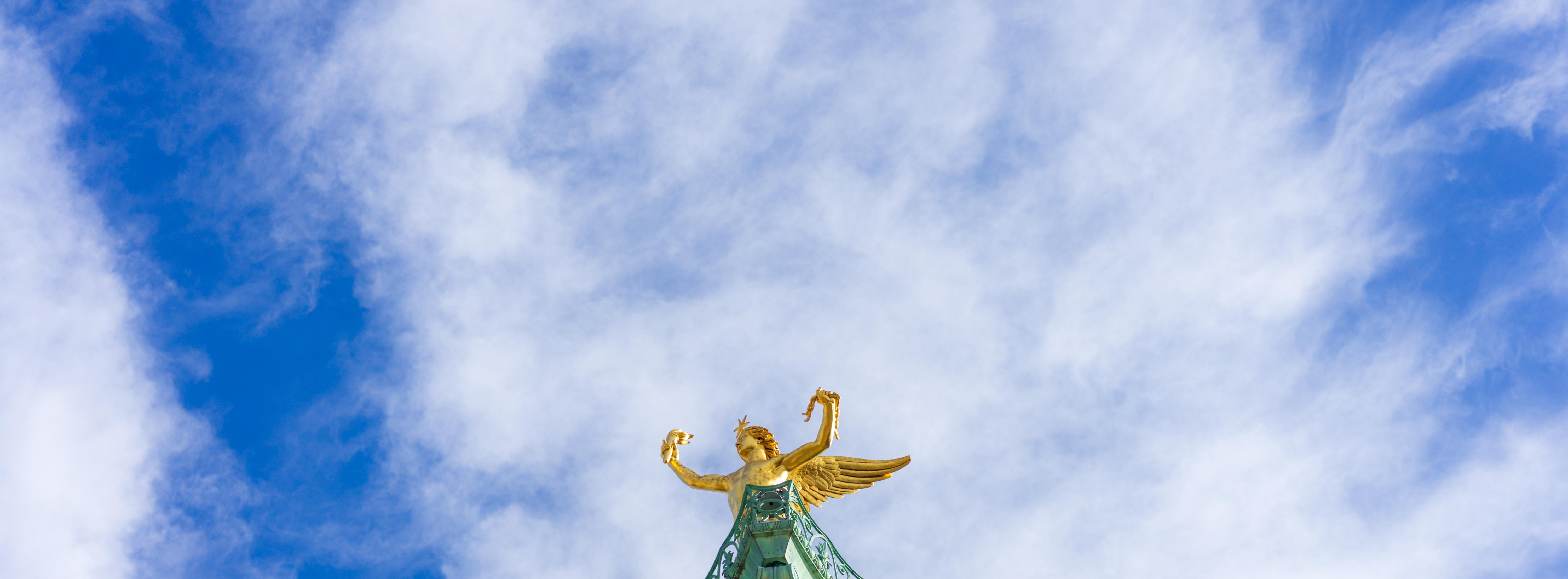Golden winged statue atop a green pedestal, set against a backdrop of bright blue sky with scattered white clouds. The statue appears to be in a triumphant pose, with arms raised.