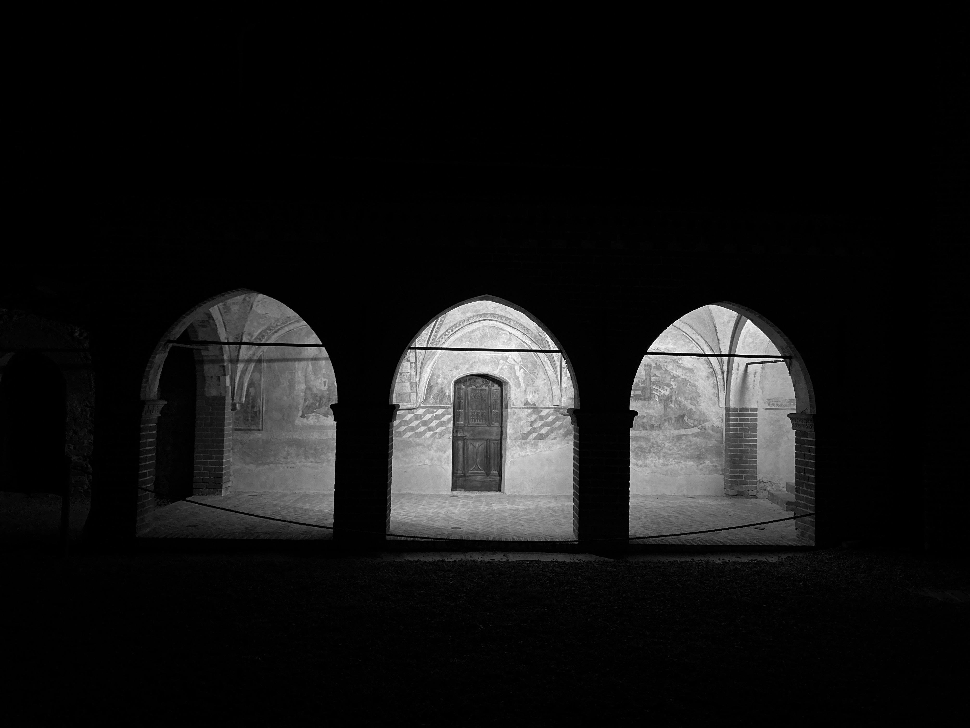 The image is a black and white photo of an architectural structure with three archways. The space behind the arches is softly lit, and there is a wooden door in the center archway. The use of light and shadow creates a dramatic effect, highlighting the arches' structure and details.