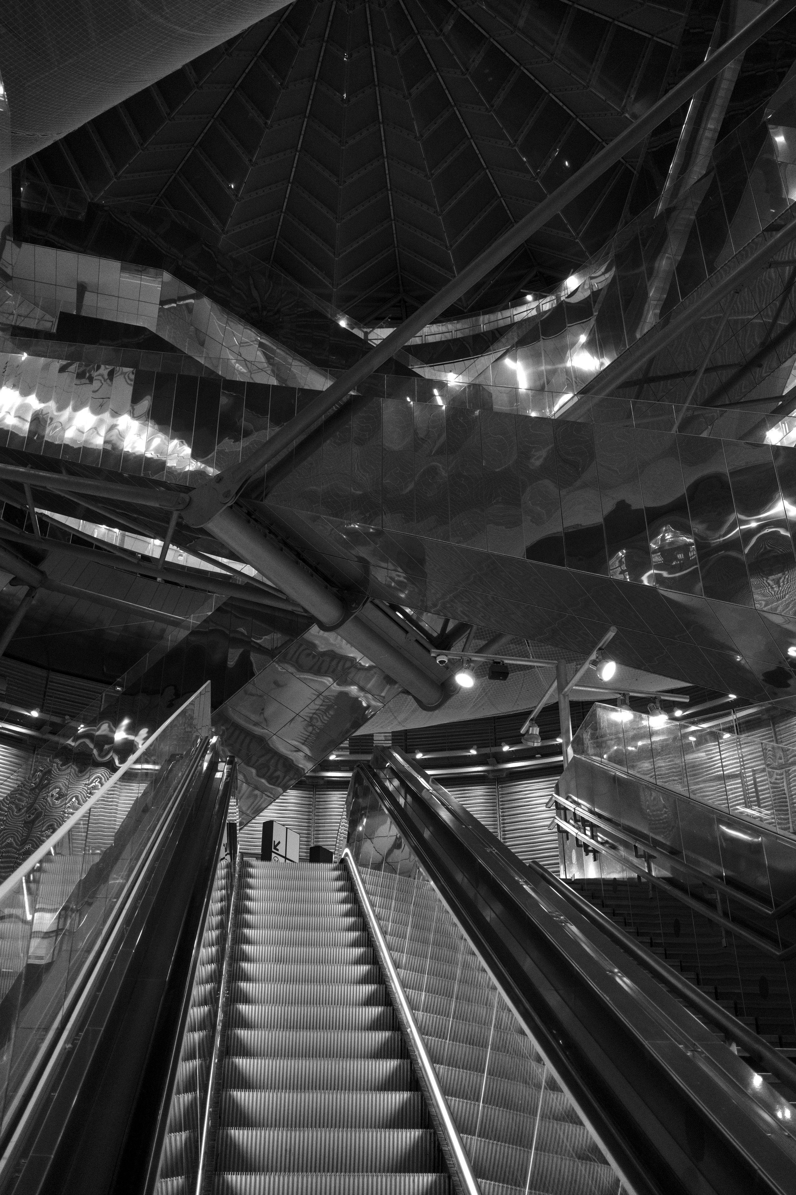 Photo verticale en noir et blanc. Un escalator qui monte depuis le premier plan et au dessus, des reflets de paroies métaliques et encore au dessus, une verrière sombre donnant sur le ciel nocturne
