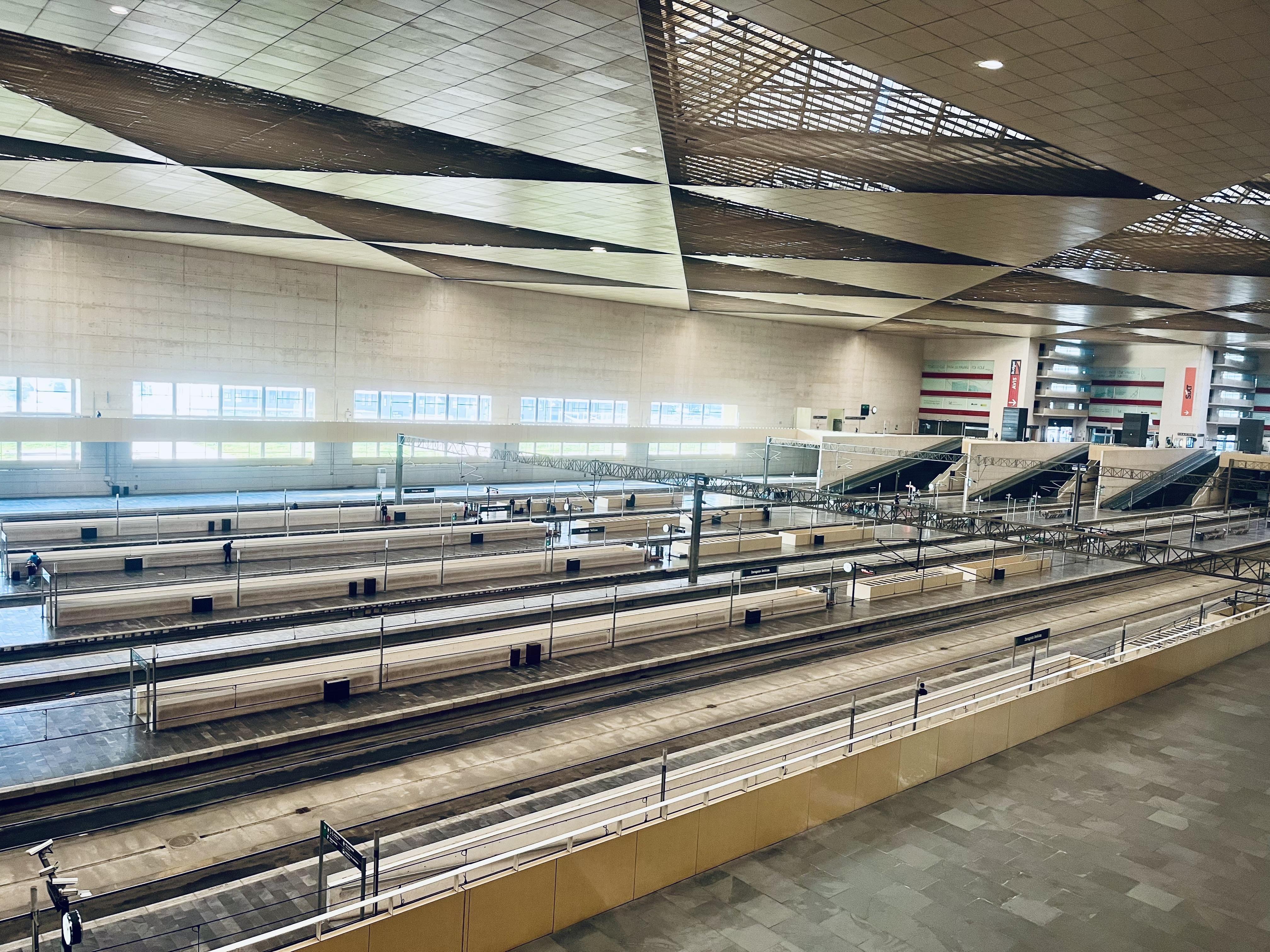 This image shows an empty, modern train station with multiple tracks and platforms. The station has a spacious interior with a high ceiling, and there are escalators leading to an upper level in the background. The architectural design features geometric patterns and ample lighting.