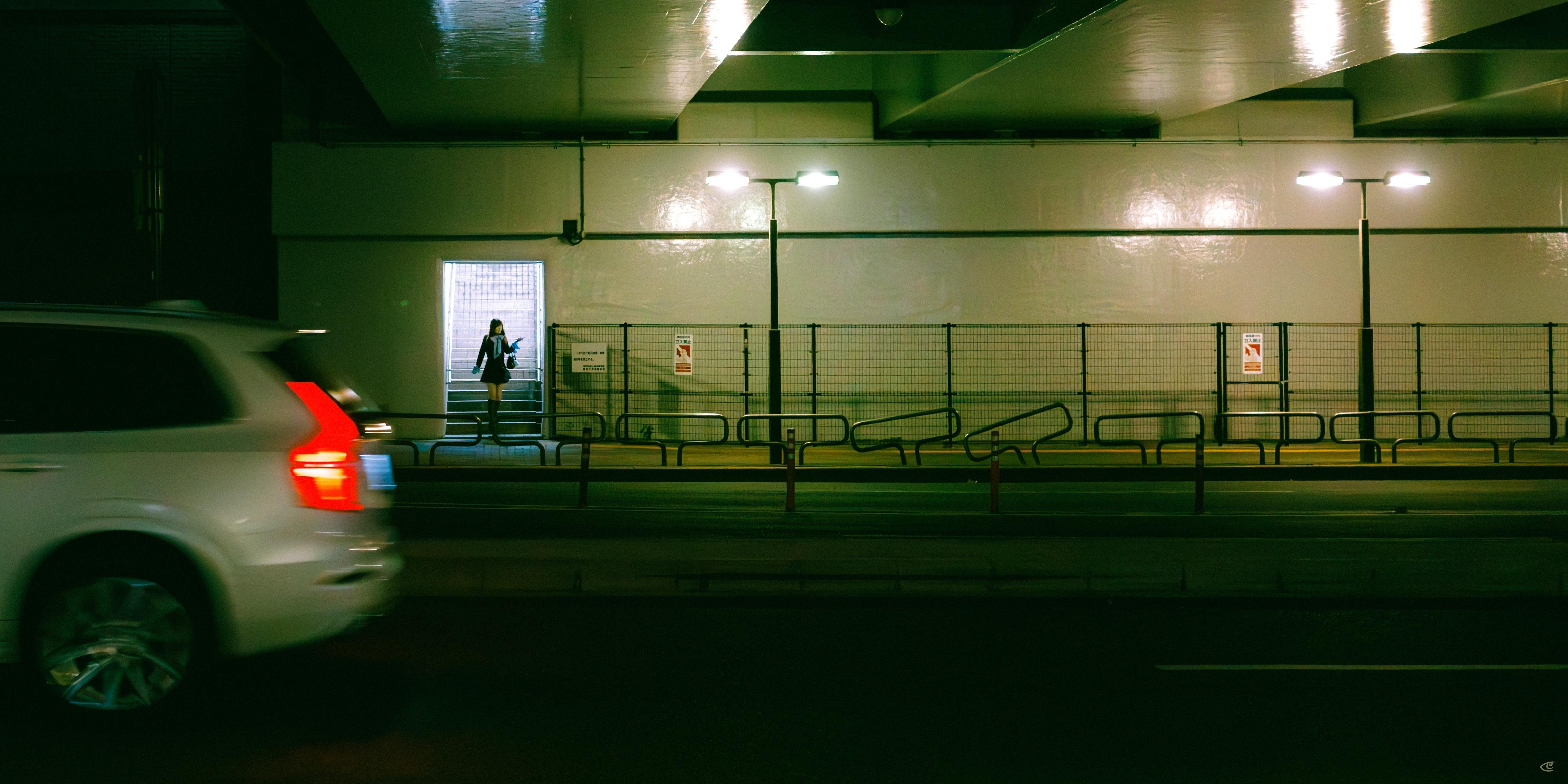Blurred white car passing in the left foreground inside a dim parking garage, with a lone person standing in a brightly lit doorway along a fenced wall under overhead lights.