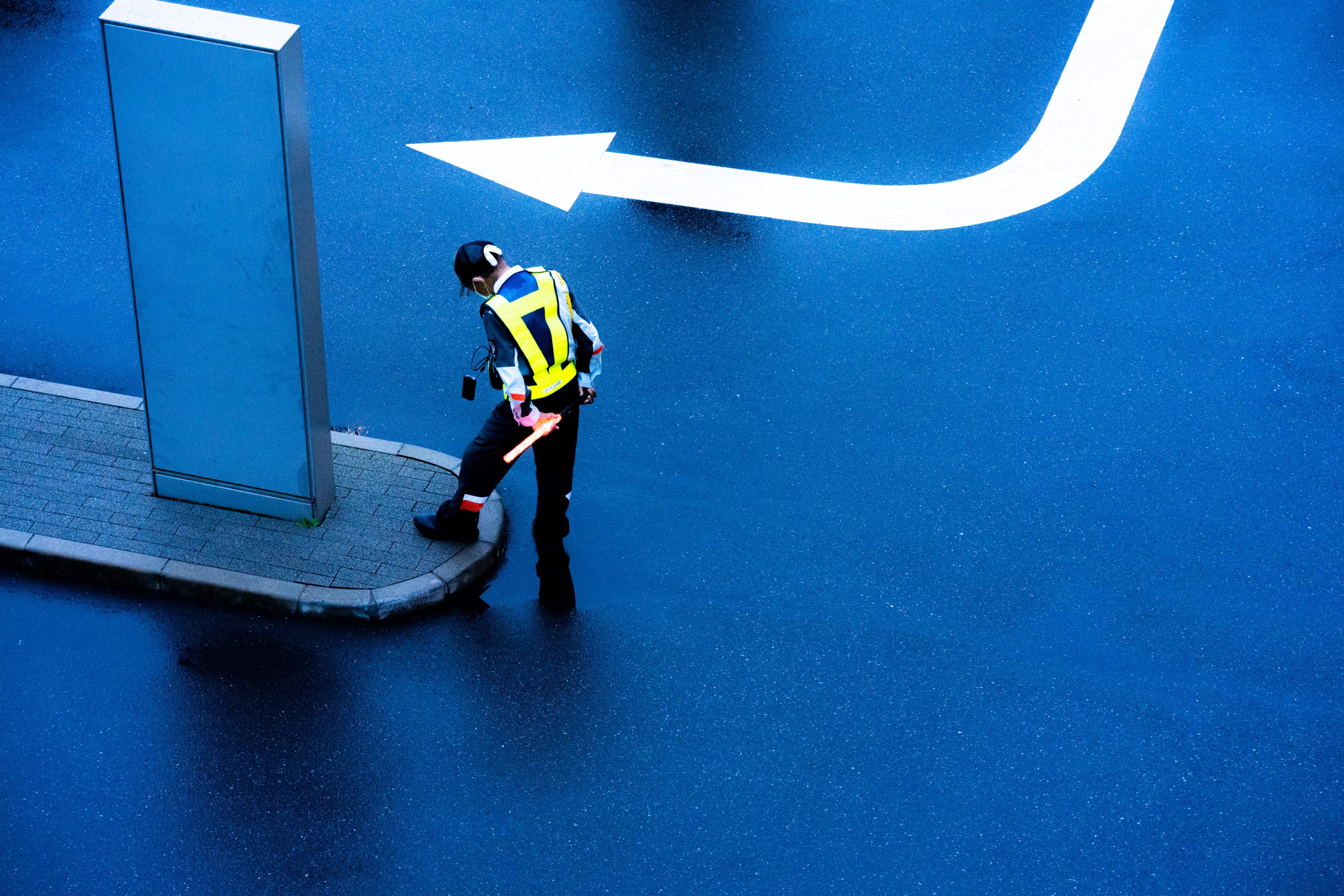 Traffic officer in high-visibility clothing stands on a wet, blue-paved street. A large white arrow painted on the road curves left. Officer carries a red baton, positioned near a narrow, raised platform.