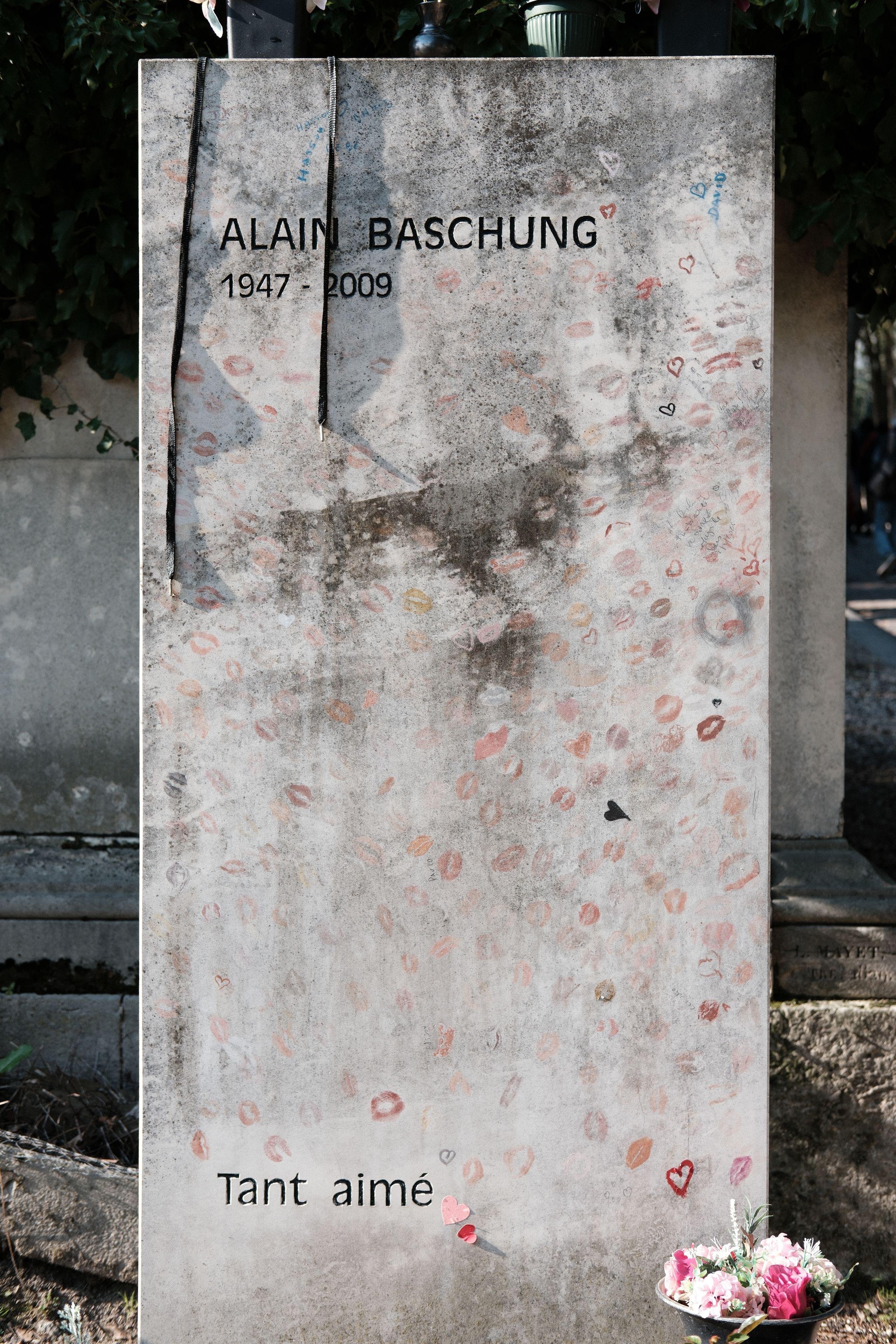 Photo verticale. Stèle de la tombe d’Alain Baschung, son nom en lettres capitales et les années 1947-2009. On y voit aussi les mots «&nbsp;Tant aimé&nbsp;» en bas à gauche. Elle est très sobre, rectangulaire, mais plein de marques de lèvres en rouge à lèvre recouvre la pierre un peu partout.