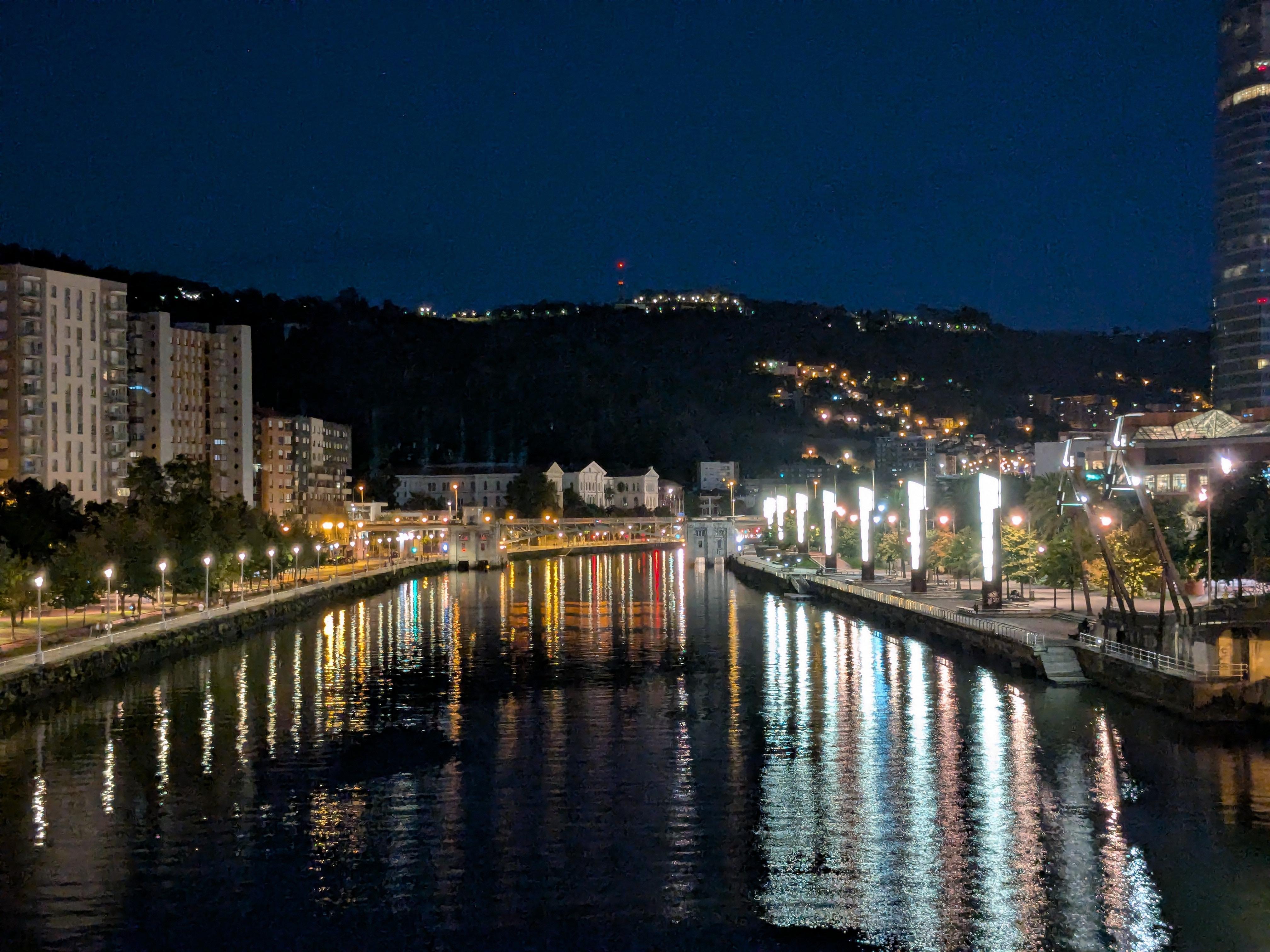 Nighttime view of a river flanked by illuminated buildings and streetlights, creating reflections on the water. On the left, tall apartment buildings line the riverbank, while to the right, a promenade with bright vertical lights stretches along the water. In the background, a hill with sparse, scattered lights and a red-lit antenna is visible.