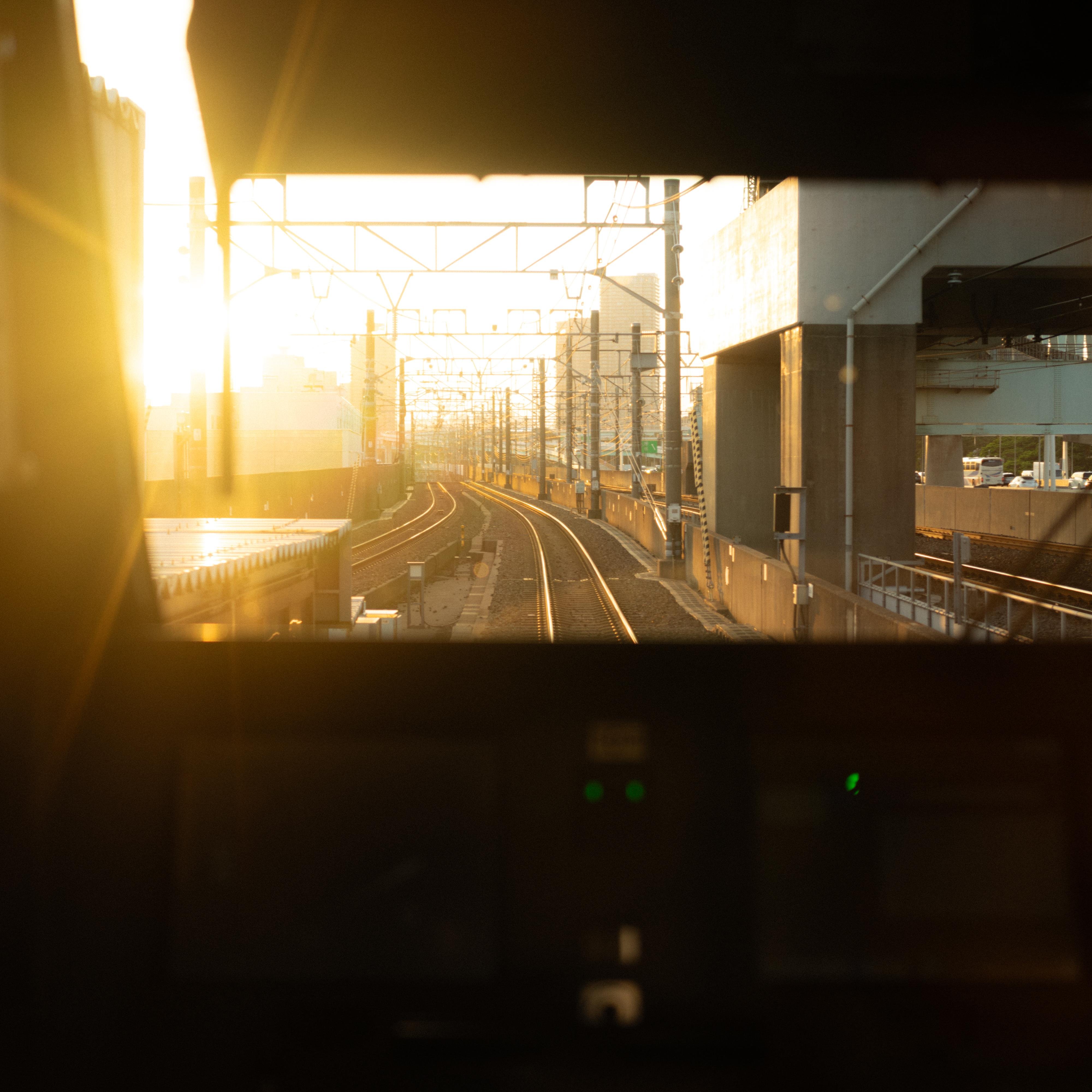 Sunlit railway tracks stretch into the distance, viewed from inside a train cab. The tracks are flanked by industrial structures and power lines. Bright sunlight creates a stark contrast and glare, casting warm hues across the scene. Control panels are partially visible in the foreground.