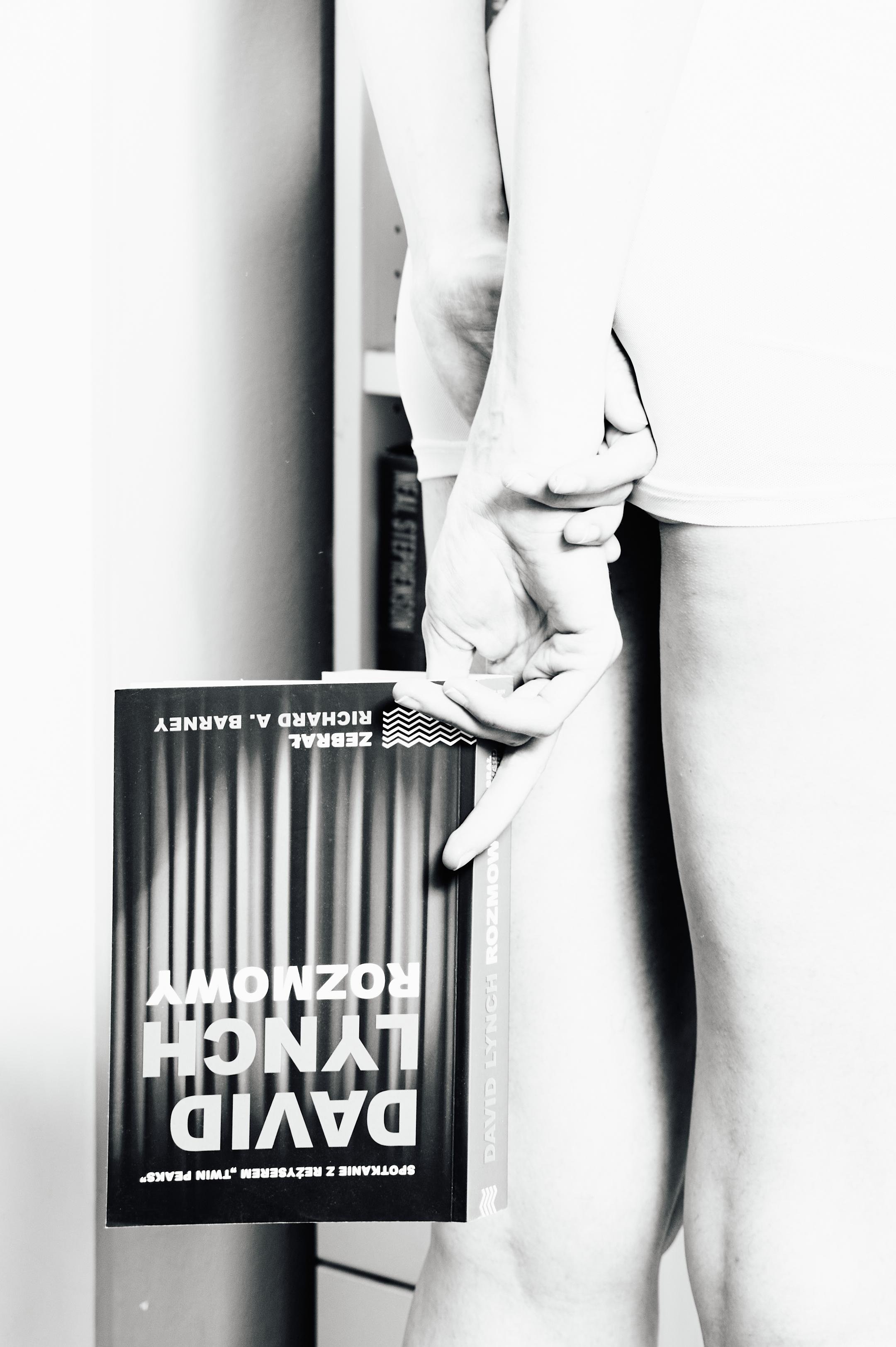 Black-and-white close-up of a person standing in shorts, holding a book upside down behind their back; the cover reads “DAVID LYNCH ROZMOWY,” with a bookshelf partially visible in the background.
