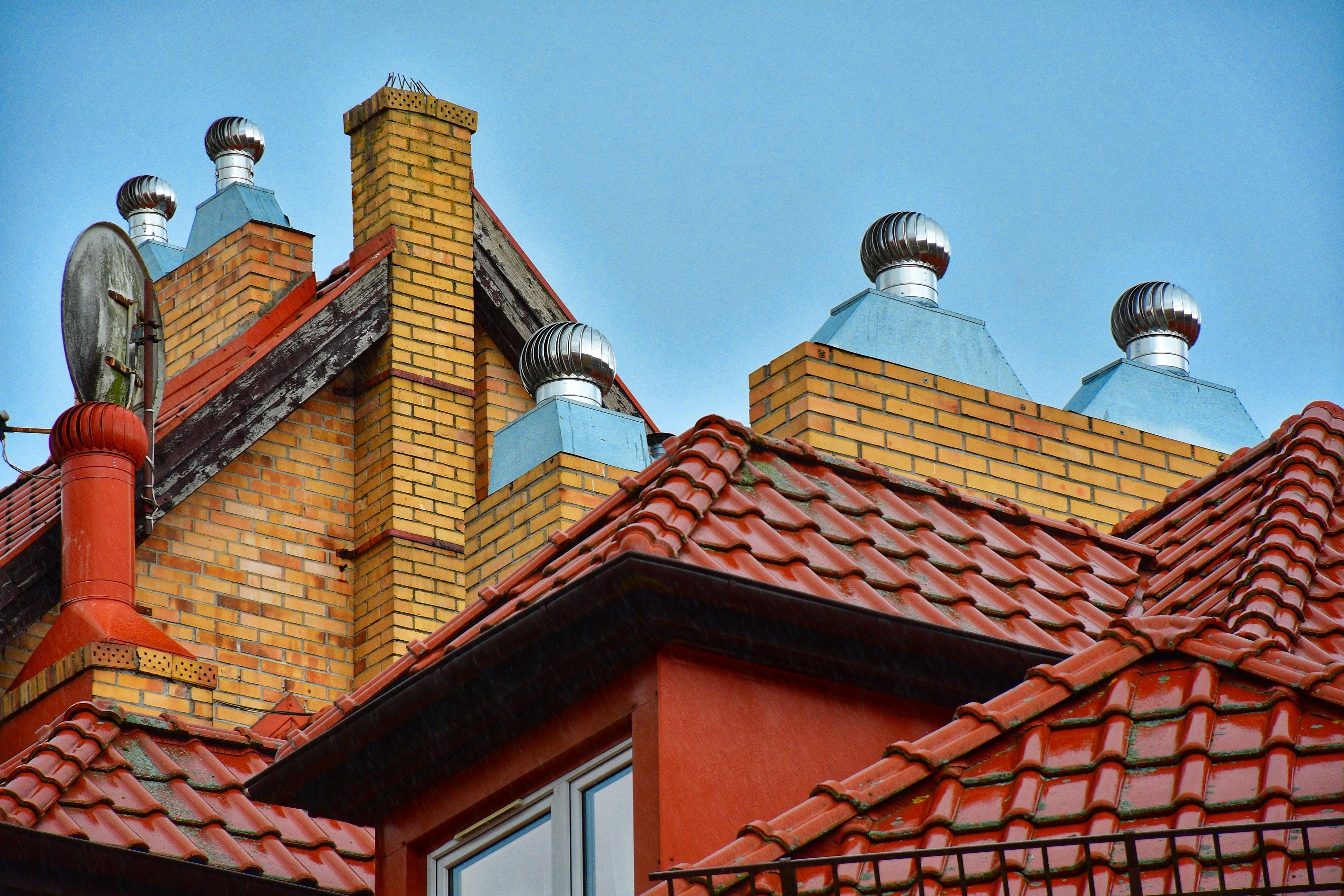 Red clay-tile rooftops with multiple yellow brick chimneys and metal roof vents, plus a satellite dish on the left, against a clear blue sky.