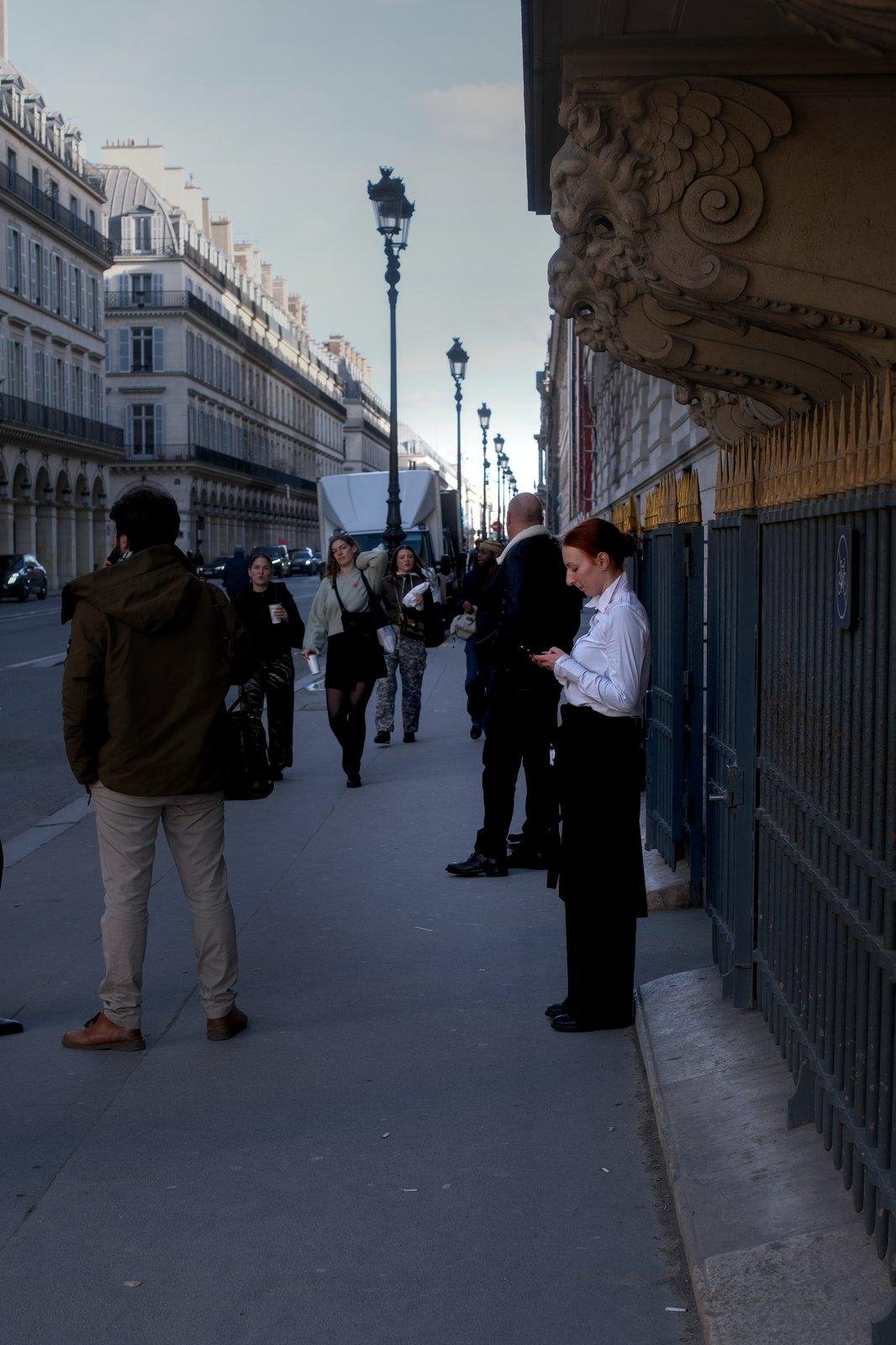 Photo verticale. Une jeune femme au cheveux colorés en rouge sombre, coiffée d’un chignon, en chemise blanche et pantalon noir, entrain de consulter son portable sur le trottoir. Des gens passent devant elle sans lui préter attention