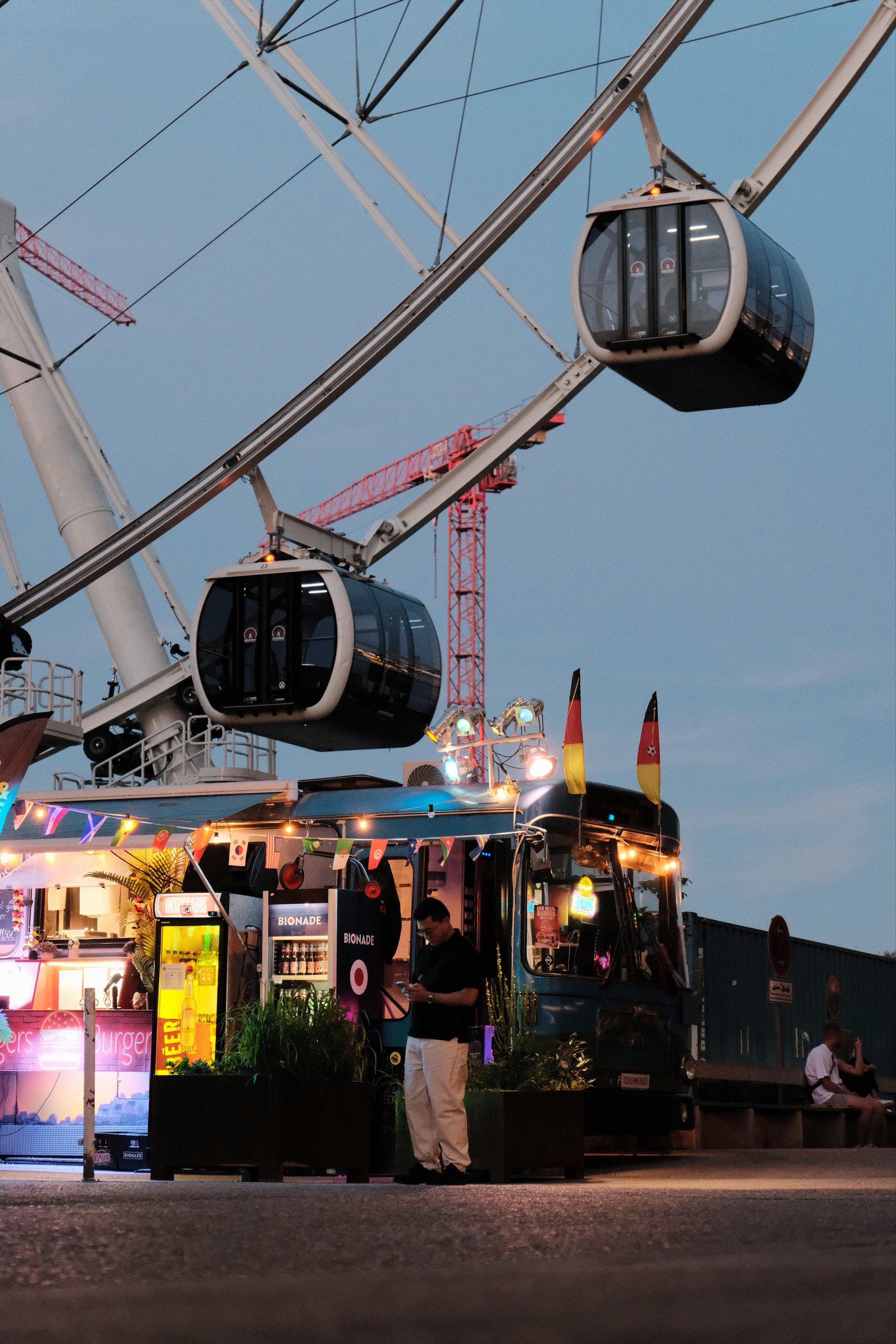 Two gondola cabins on a Ferris wheel above a brightly and colourfully lit food truck, with a man standing nearby.