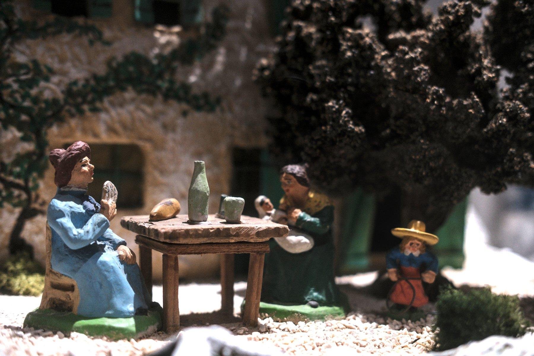 Photo horizontale. Un morceau de diorama en santon, représentant une femme attablée à l’extérieur, en train de manger quelque chose. Une miche de pain, une bouteille et un verre sont sur la table. À l’opposé se trouve une autre femme qui tiens un bébé dans ses bras. Enfin, un enfant avec un chapeau jaune joue sous un grand arbre
