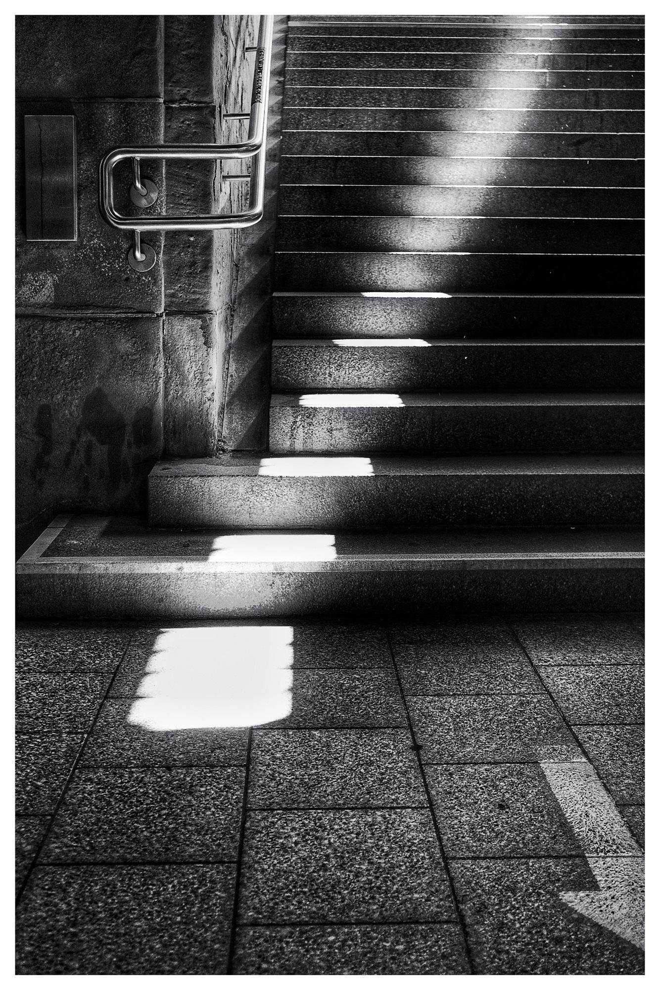 Black-and-white view of a stone stairway rising to the right, with a metal handrail on the left wall and rectangular patches of sunlight falling across the steps and tiled floor at the base.