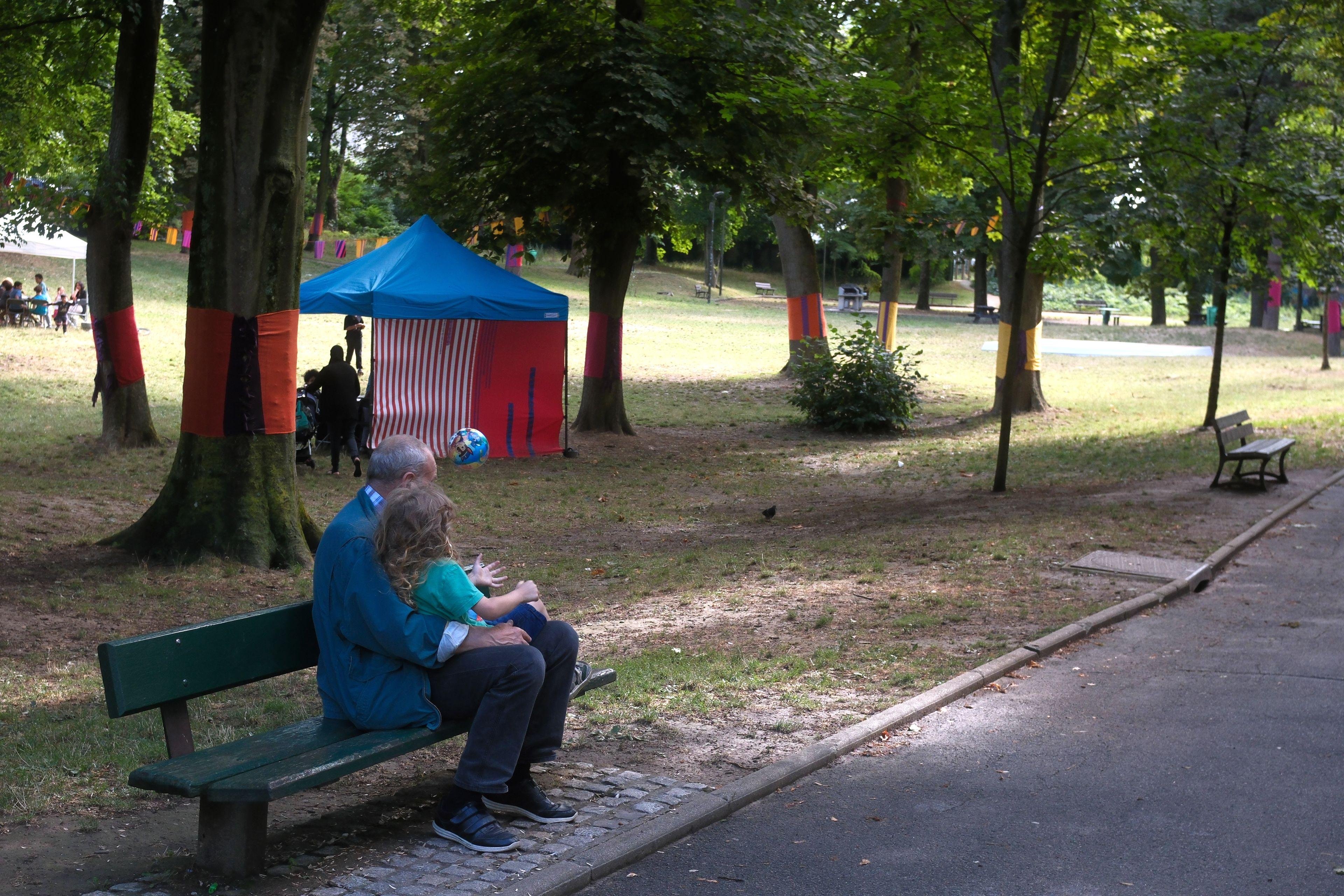 A photo of the back an old man with a young girl on his lap sitting on a bench in a park. The girl throw a ball caught mid air by the camera.
