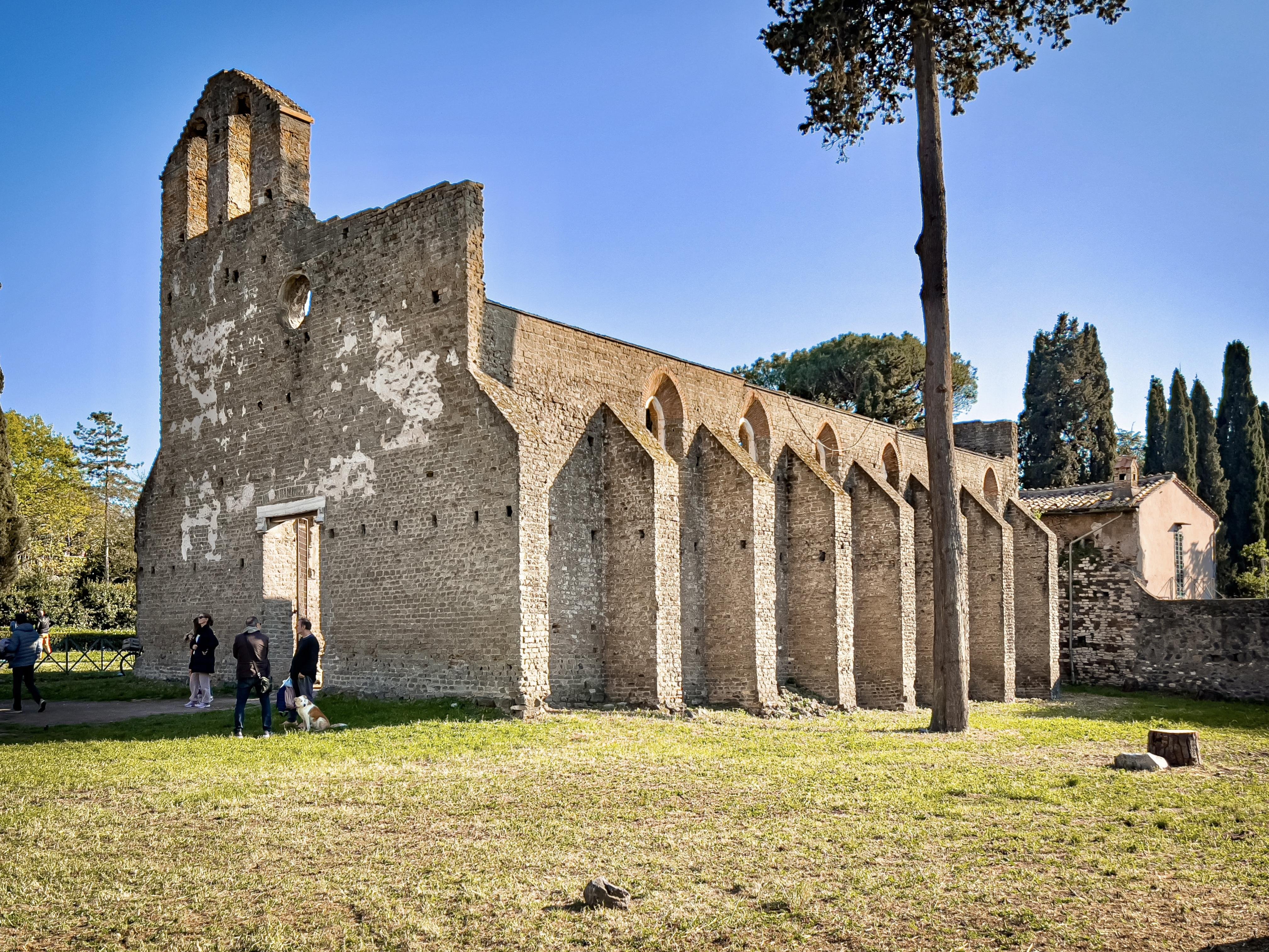This image shows the ruins of an old brick building, likely a church or abbey, characterized by its tall walls and arched window designs. The structure has a rustic appearance with some weathered areas. There are a few people and a dog in the foreground, and the setting is a grassy area with trees, suggesting a park or historical site.