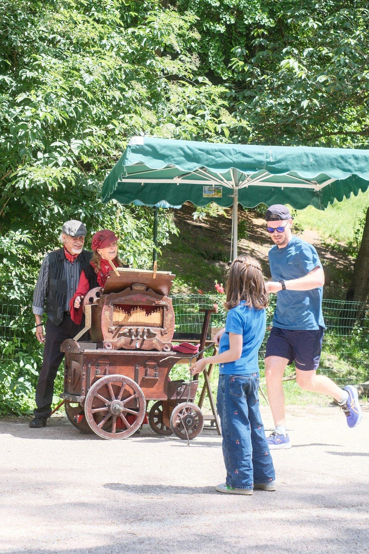 Photo verticale. Au premier plan, une fillette châtain de dos, en jean bleu et t-shirt bleu, regardant attentivement un orgue de barbarie opéré par une dame avec une casquette rouge, accompagnée de son mari, en casquette grise