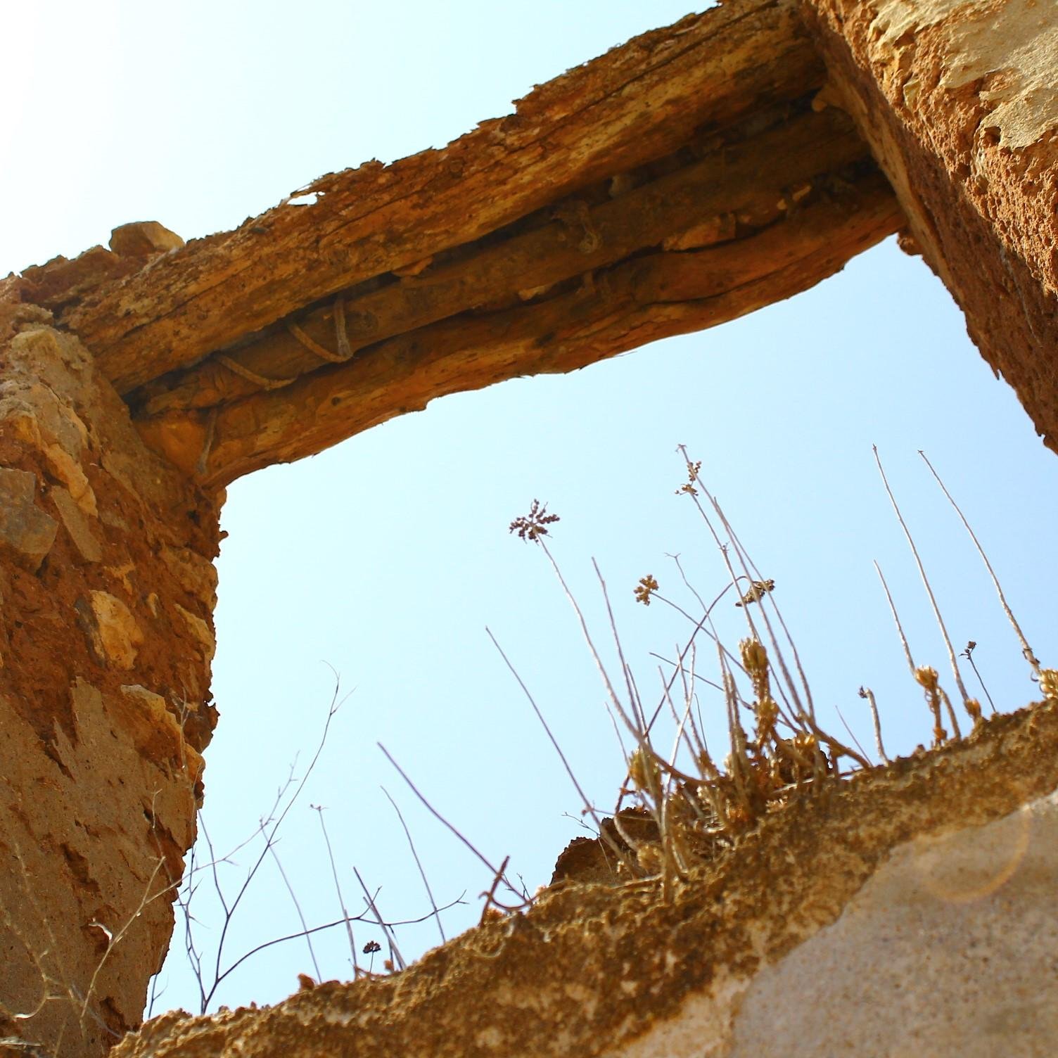Fotografía en contrapicado en la que se aprecian las ruinas de un muro antiguo en el que se encuentra el hueco de lo que alguna vez fue una puerta o una ventana. Se aprecian restos de madera y soga. Por abajo, hay maleza silvestre. Los restos son de colores marrones, amarillentos y anaranjados, en contraste con el azul claro del cielo.