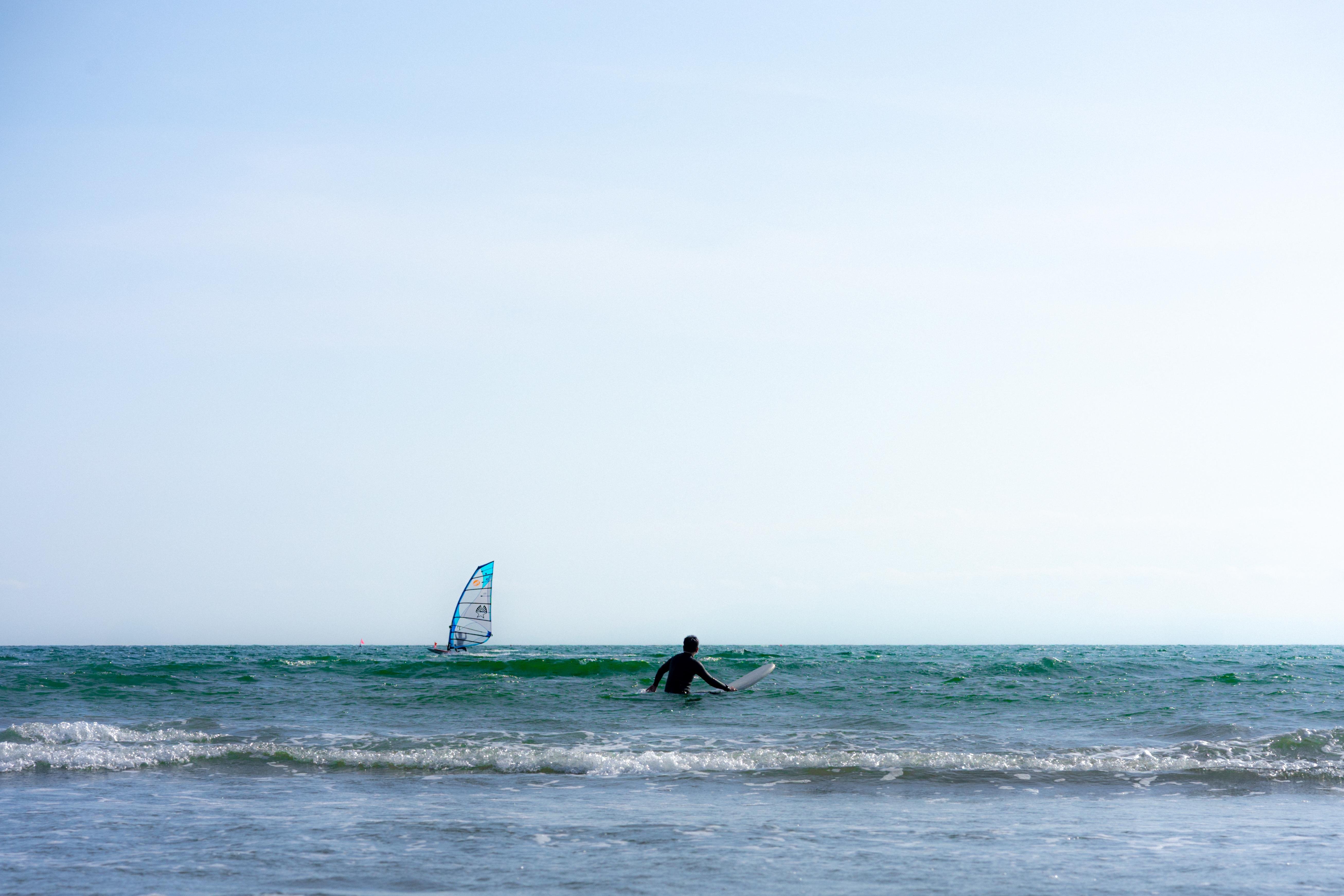 Person in a wetsuit standing in waist-deep water with a surfboard, facing the ocean. A windsurfer with a colorful sail is visible in the background, gliding over the waves. The sky is clear and blue, and gentle waves are breaking on the shore.