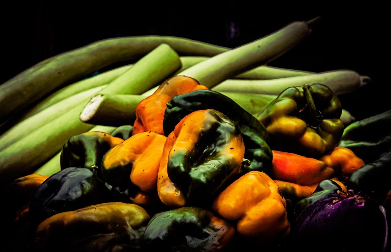 Pile of mixed vegetables, with green and orange bell peppers and small orange peppers in the foreground, long pale-green gourds or squash stacked behind them against a dark background.