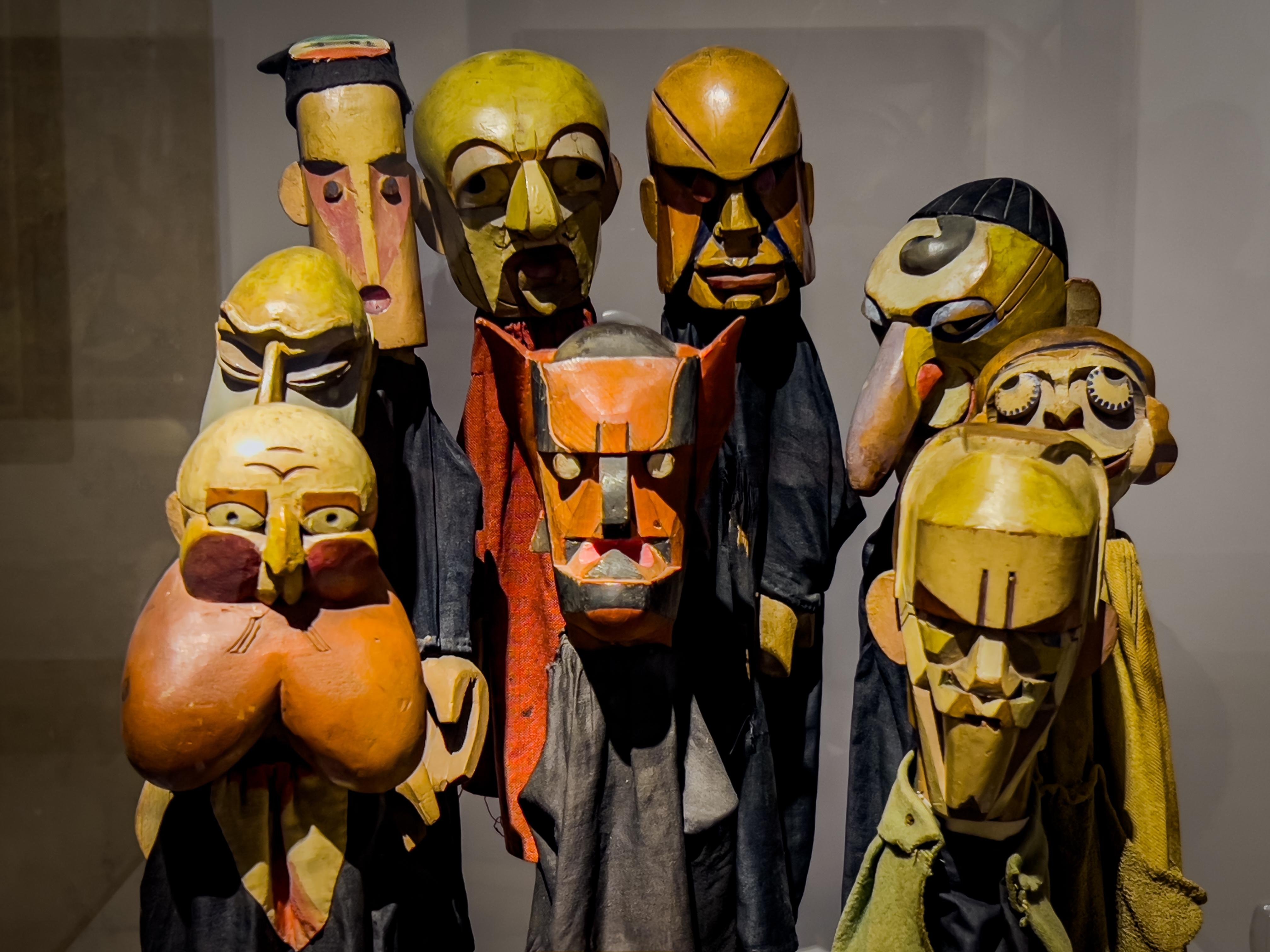 Raccolta di maschere stilizzate e astratte o figure in legno intagliato. Ogni figura presenta tratti del viso esagerati e distinte forme geometriche, mostrando una varietà di espressioni. I colori utilizzati sono prevalentemente toni della terra, con alcuni rossi, gialli e neri. La disposizione suggerisce che potrebbero far parte di una mostra d'arte o di un'installazione.