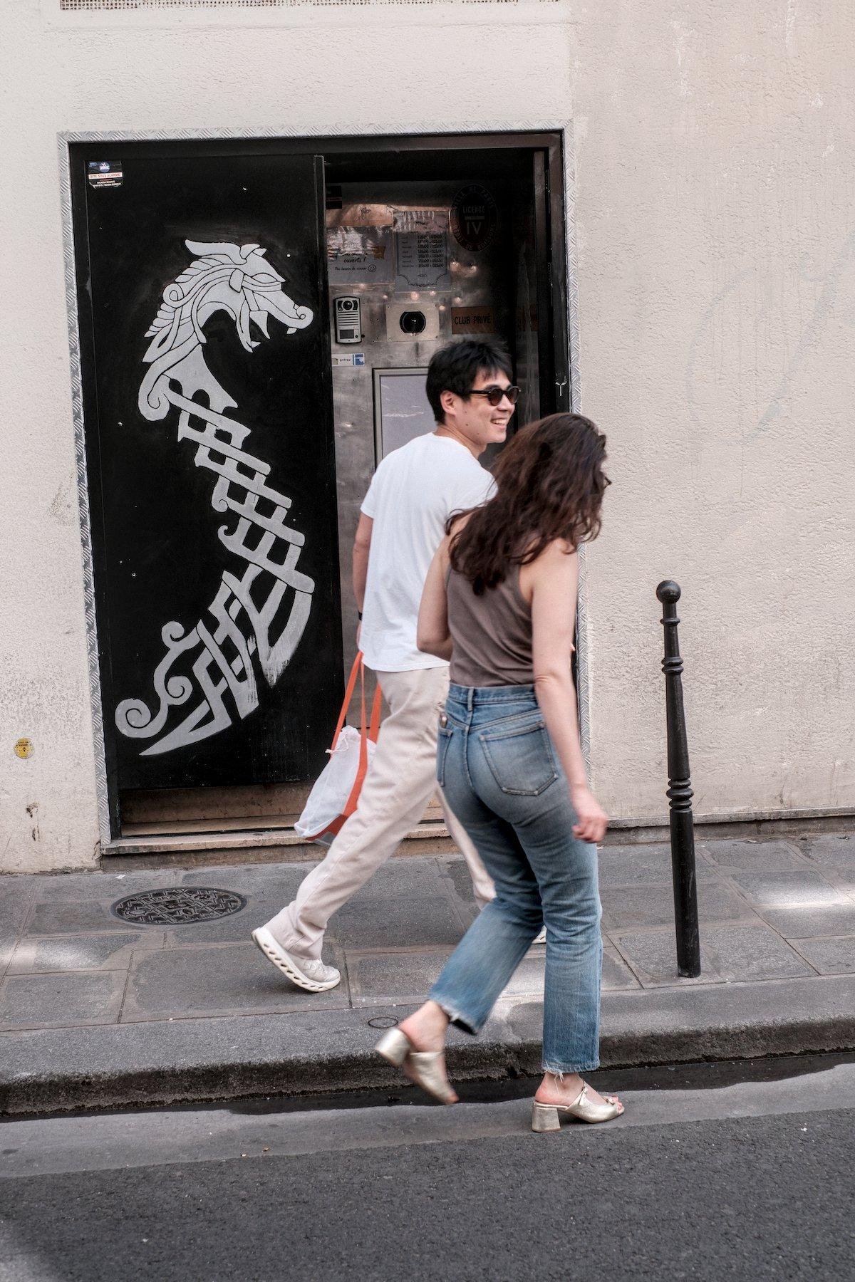 Photo verticale. Une double porte noire avec un dragon blanc stylisé peint sur celle de gauche, dans un style viking. La porte de droite est ouverte découvrant une autre porte en métal avec une caméra interphone. 2 personnes passent devant celle-ci