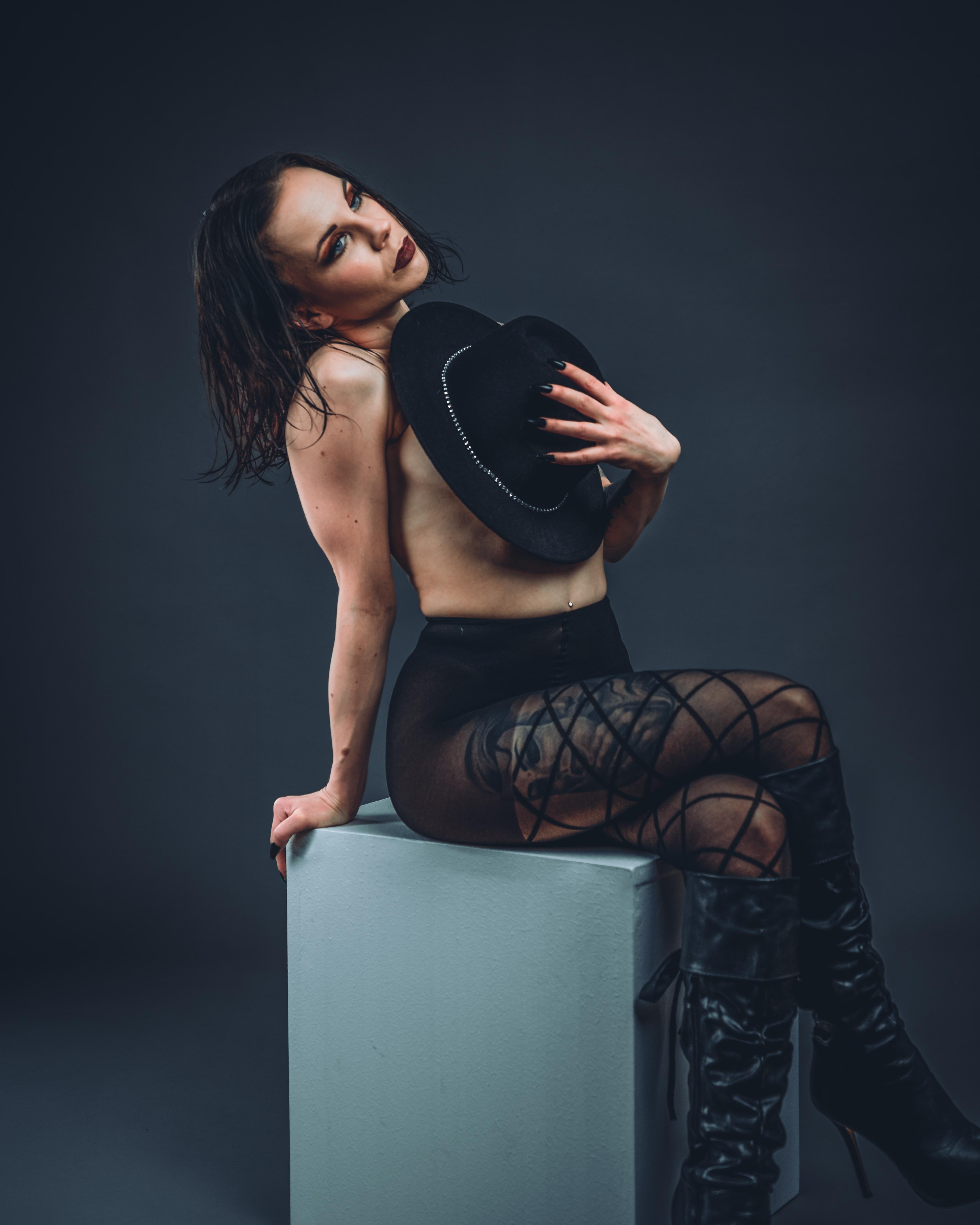 Person sitting on a gray block against a dark background, holding a black hat partially covering their chest. They wear black sheer tights with a lattice pattern and knee-high black boots. Head tilted slightly back with wet hair and dramatic makeup.