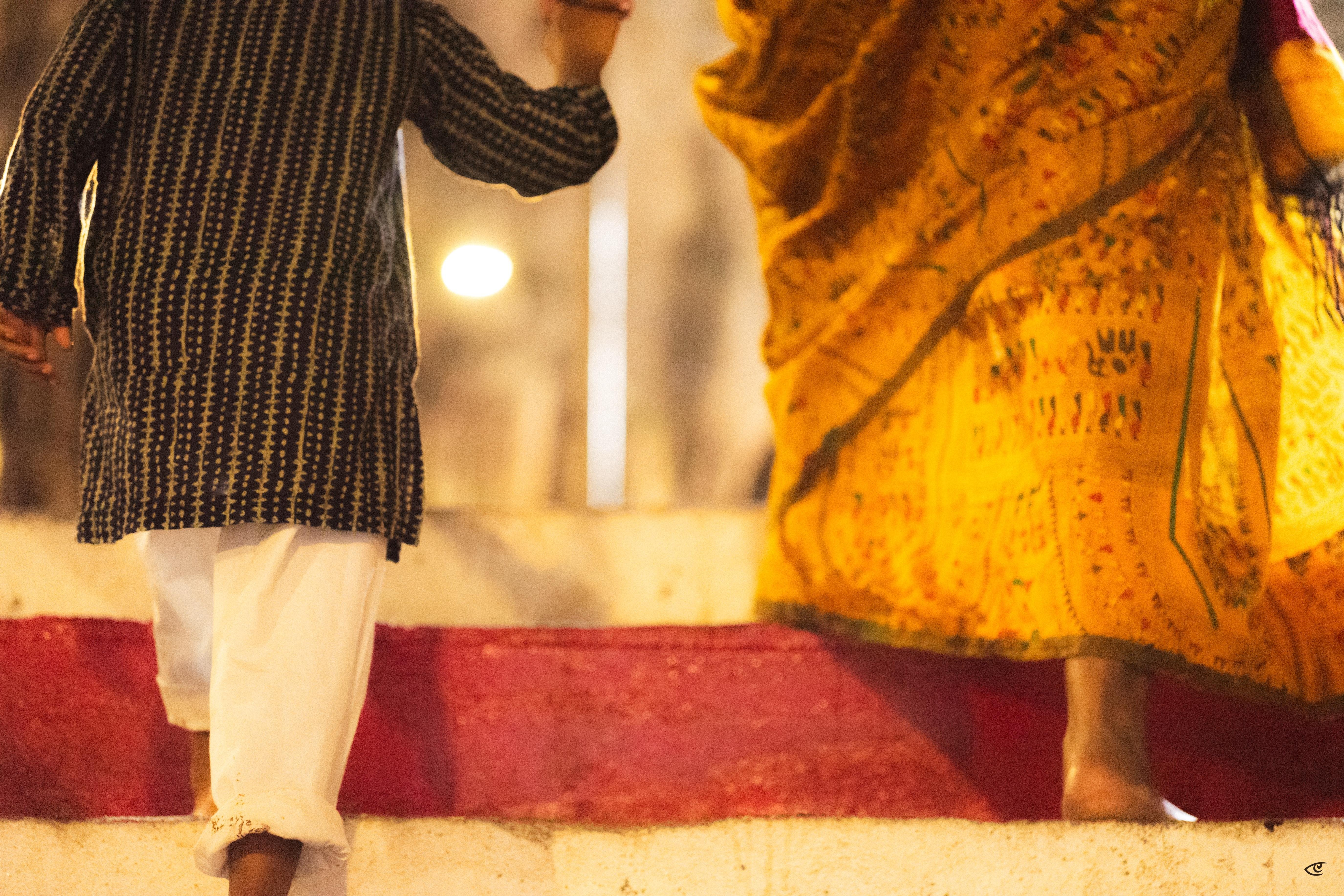 Lower-body close-up of two people stepping down a curb, one wearing a dark patterned tunic with white pants and the other wearing a flowing yellow patterned skirt, with warm blurred streetlights in the background.