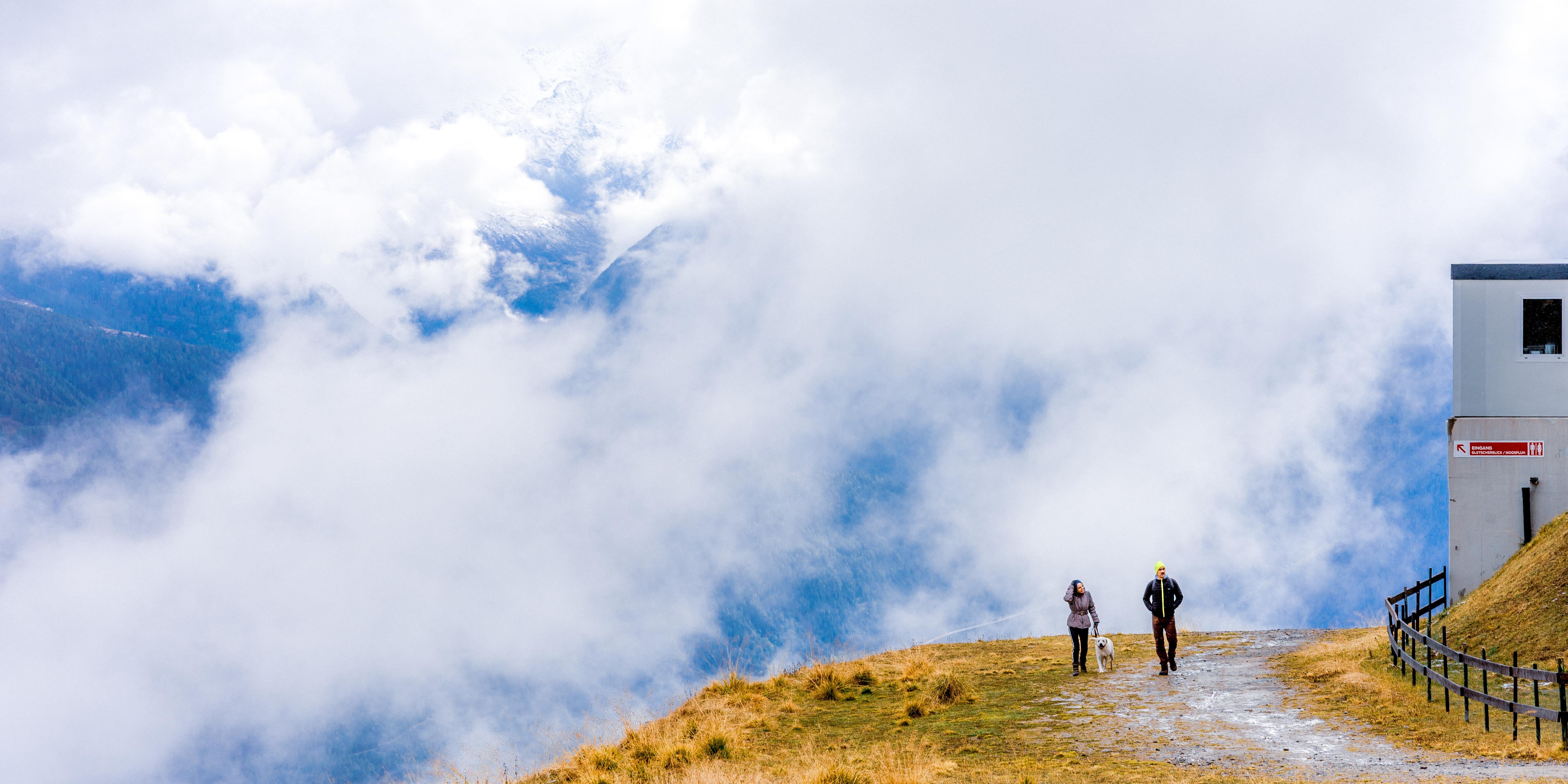 Two people walking along a gravel path on a grassy hillside surrounded by clouds. A white building with a red sign is on the right. The background features a cloud-covered mountainous landscape.