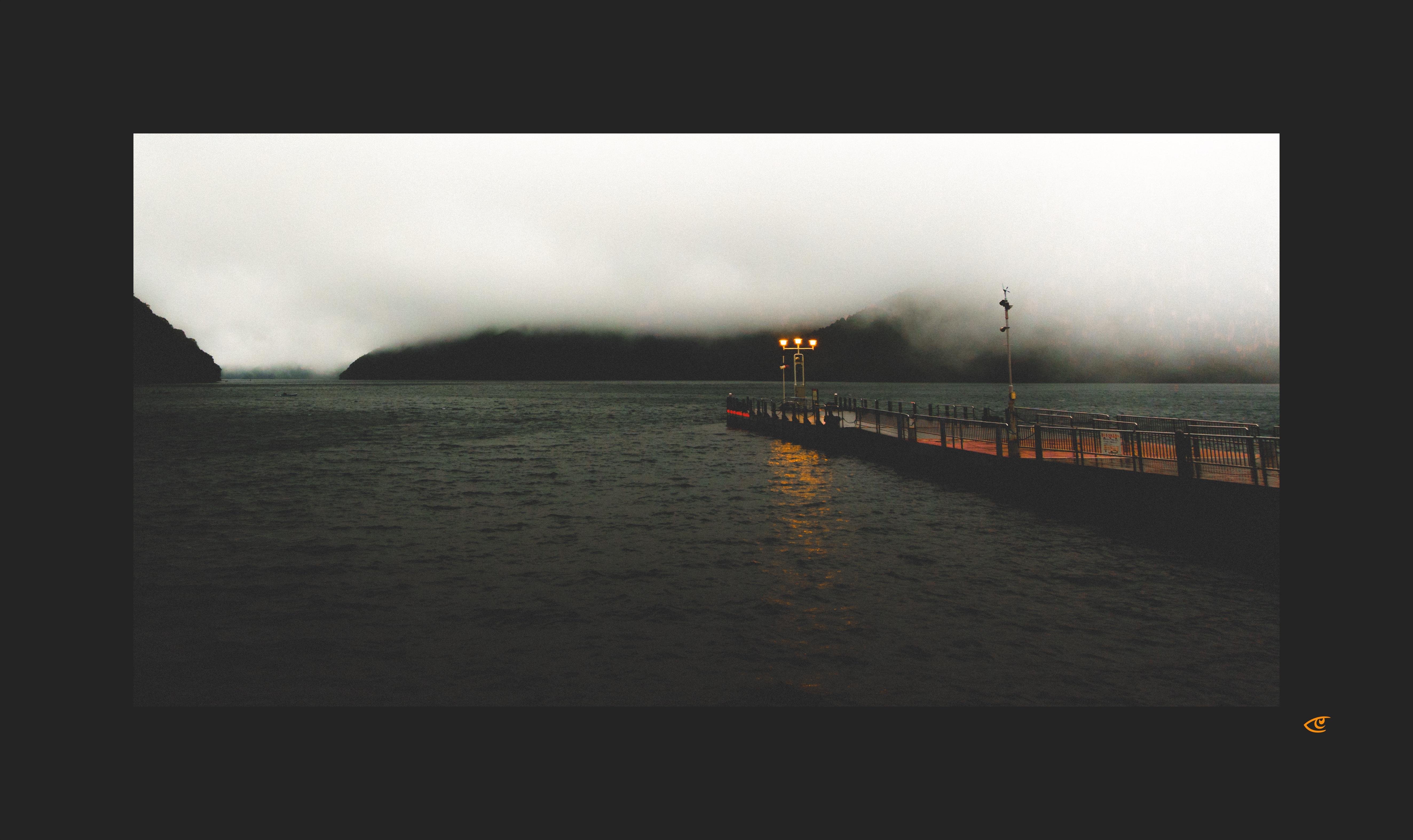 Long pier with metal railings extending over dark water toward mist-covered mountains, with two lit lamps near the end reflecting on the water under an overcast sky.