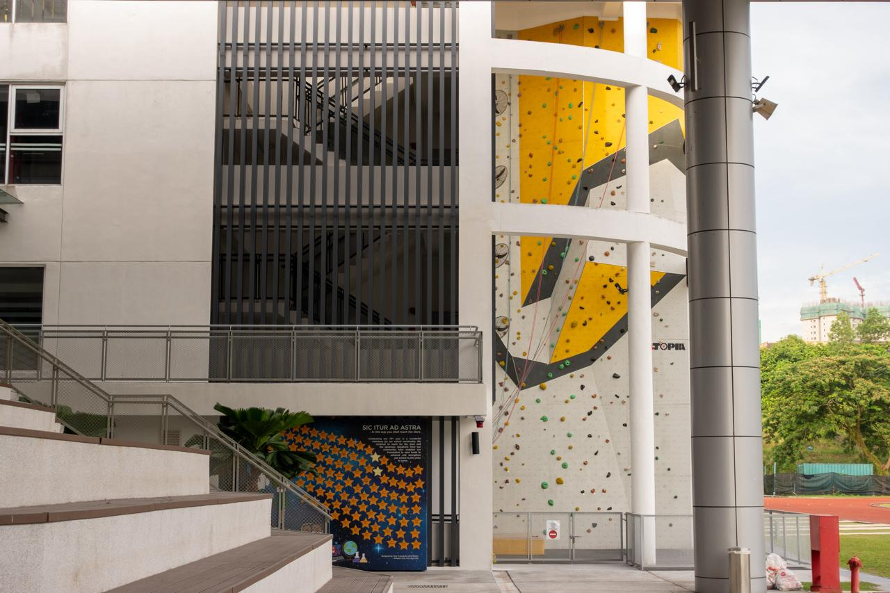Outdoor rock climbing wall with yellow and black geometric patterns, featuring handholds in various colors. Adjacent to the wall, a modern building with a stairwell enclosed by vertical metal bars. A stairway with wooden steps leads down to the area. A sign with text and star graphics is displayed near the base. A running track and greenery are seen in the background.