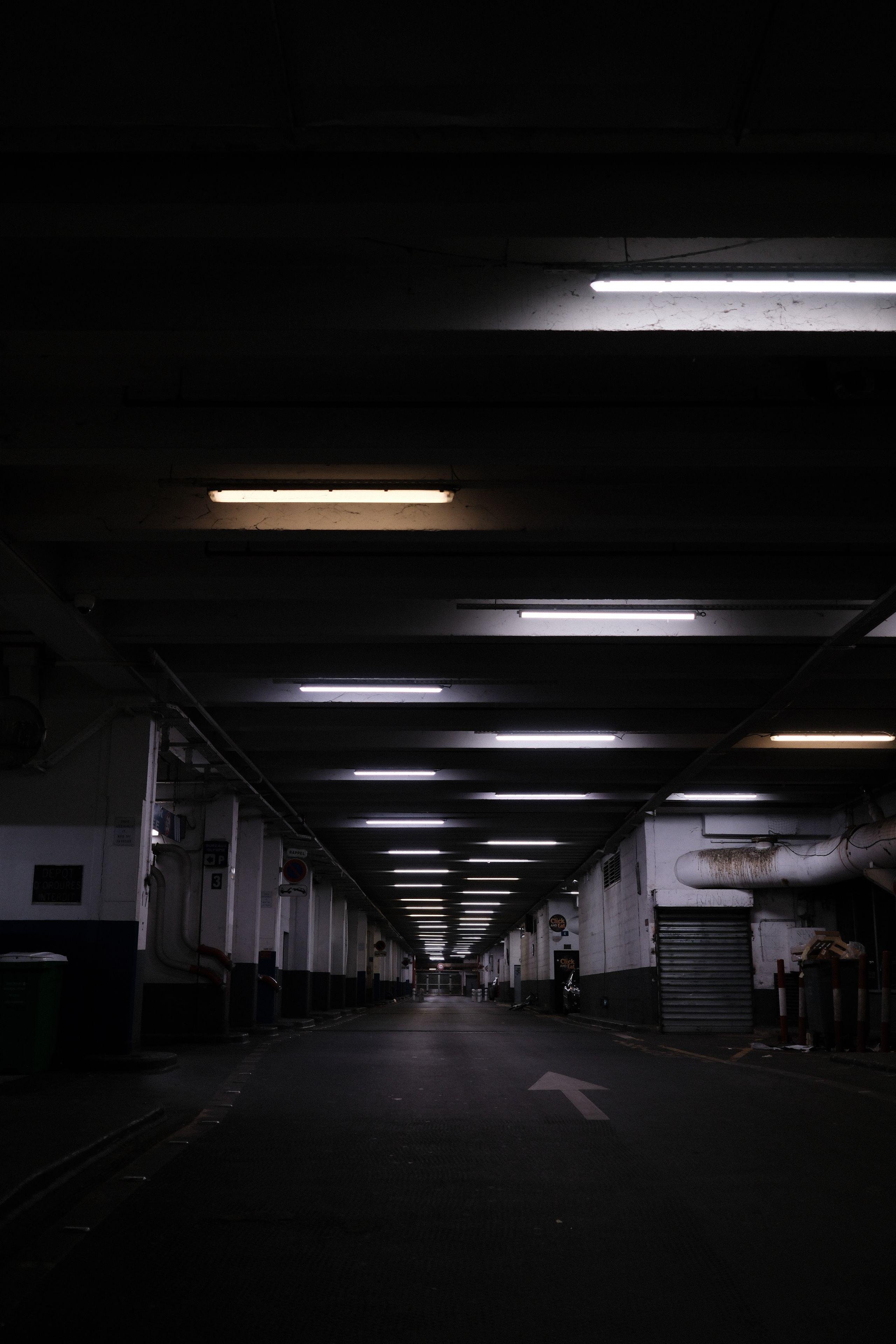Photo format portrait d’une route souterraine. On voit du côté gauche des poteaux se répétant vers le point de fuite, pareil pour le haut avec des néons blafards en quinconce, et à droite, on voit un box fermé d’un rideau de fer gris avec au dessus un gros tuyau blanc sortant du box, mais tout salis d’une coulée noire/marron sur le début. Puis toujours à droite, d’autre box se répètent