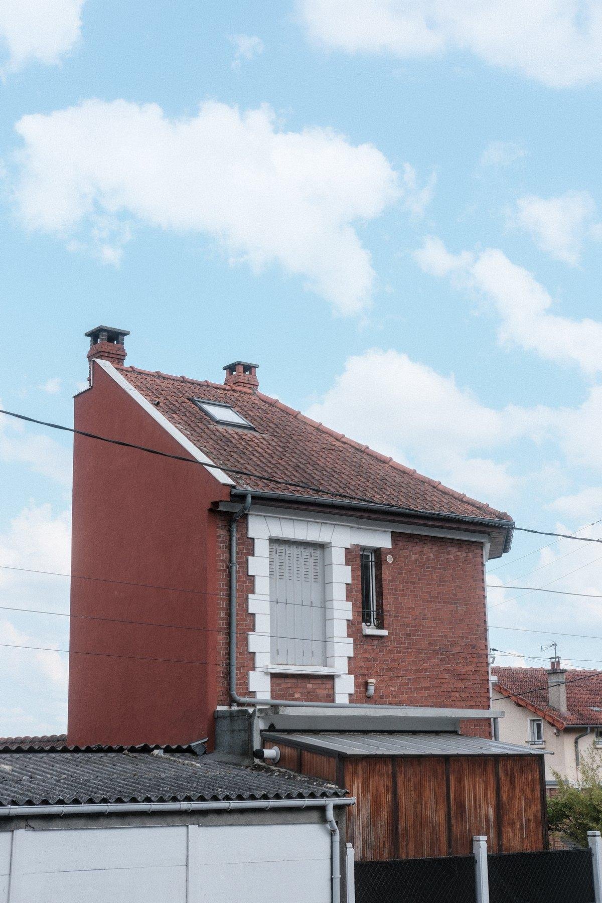 Photo verticale. Une maison en brique rouge, avec une extension en bois usé. Sa forme fait penser à un quart de maison