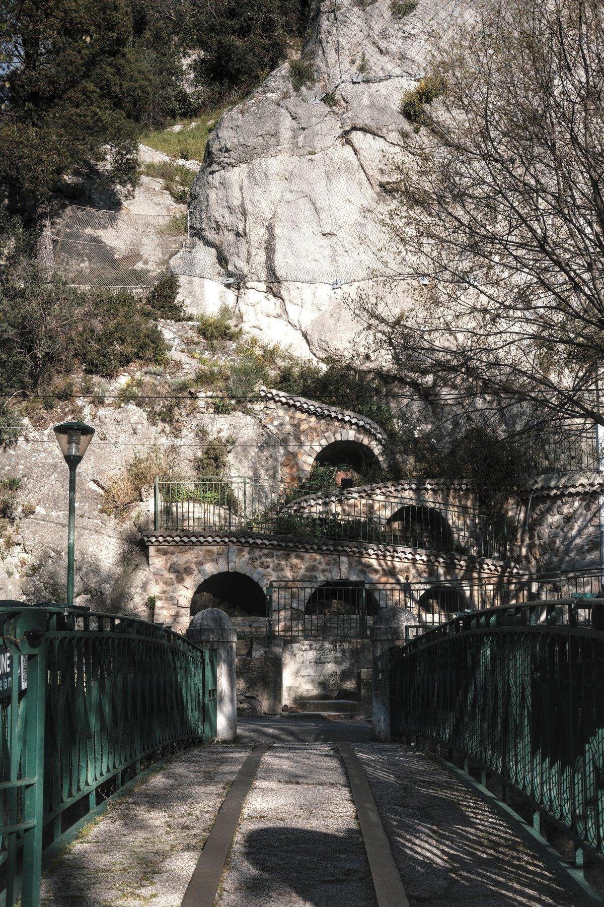 Photo verticale. Depuis l’extrémité d’un pont piéton, on voit l’autre rive, menant à une falaise dans laquelle on distingue un escalier qui zig zag sur des murs en pierre avec des arches