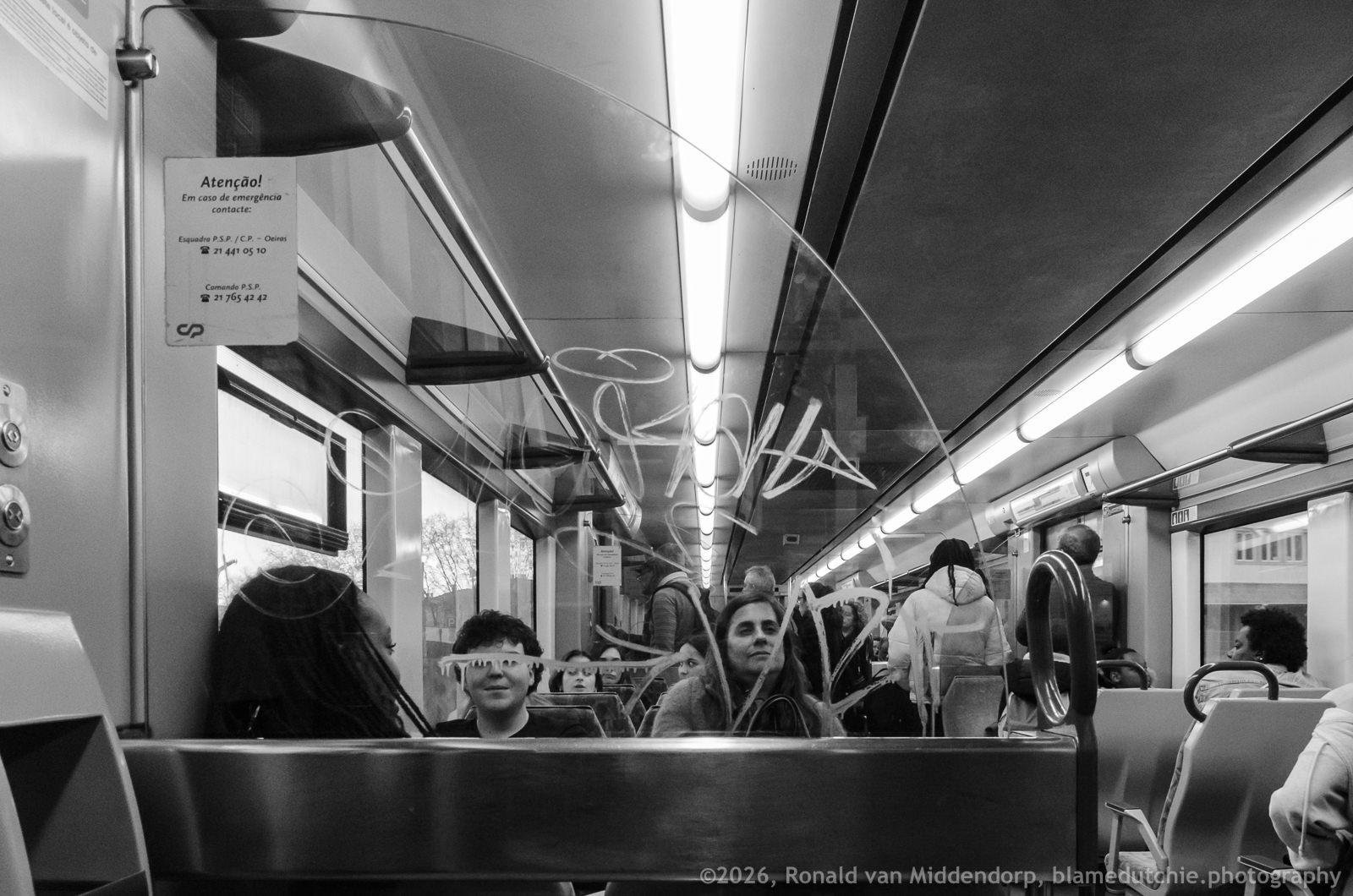 Black-and-white view down the aisle of a train carriage with fluorescent ceiling lights, passengers seated and standing, and a glass partition covered in graffiti in the foreground; an information notice is posted on the left wall above the seats.