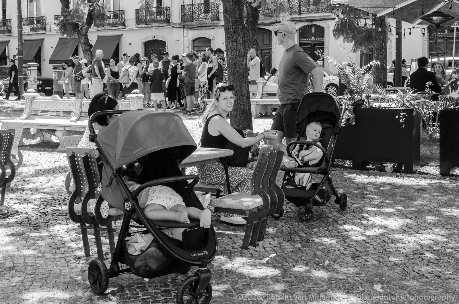 Black-and-white photo of a cobblestone plaza with café tables and chairs under trees; a stroller in the foreground holds a child lying down, while a woman sits at a table beside a second stroller with a child and a standing man, with a crowd and storefronts in the background.