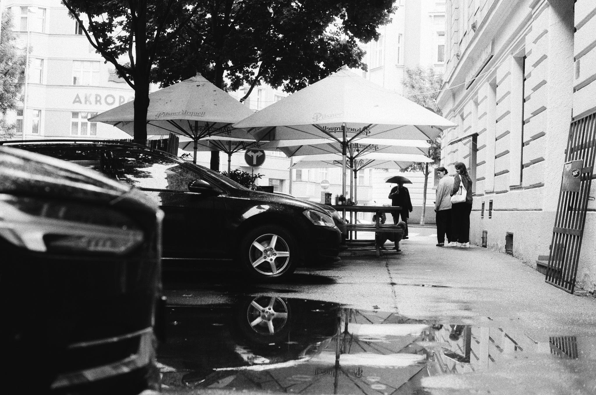 Black-and-white street scene with parked cars along the left, a puddle in the foreground reflecting a car wheel and umbrellas, and two large patio umbrellas shading a bench in the middle distance; two people stand near the building wall on the right.