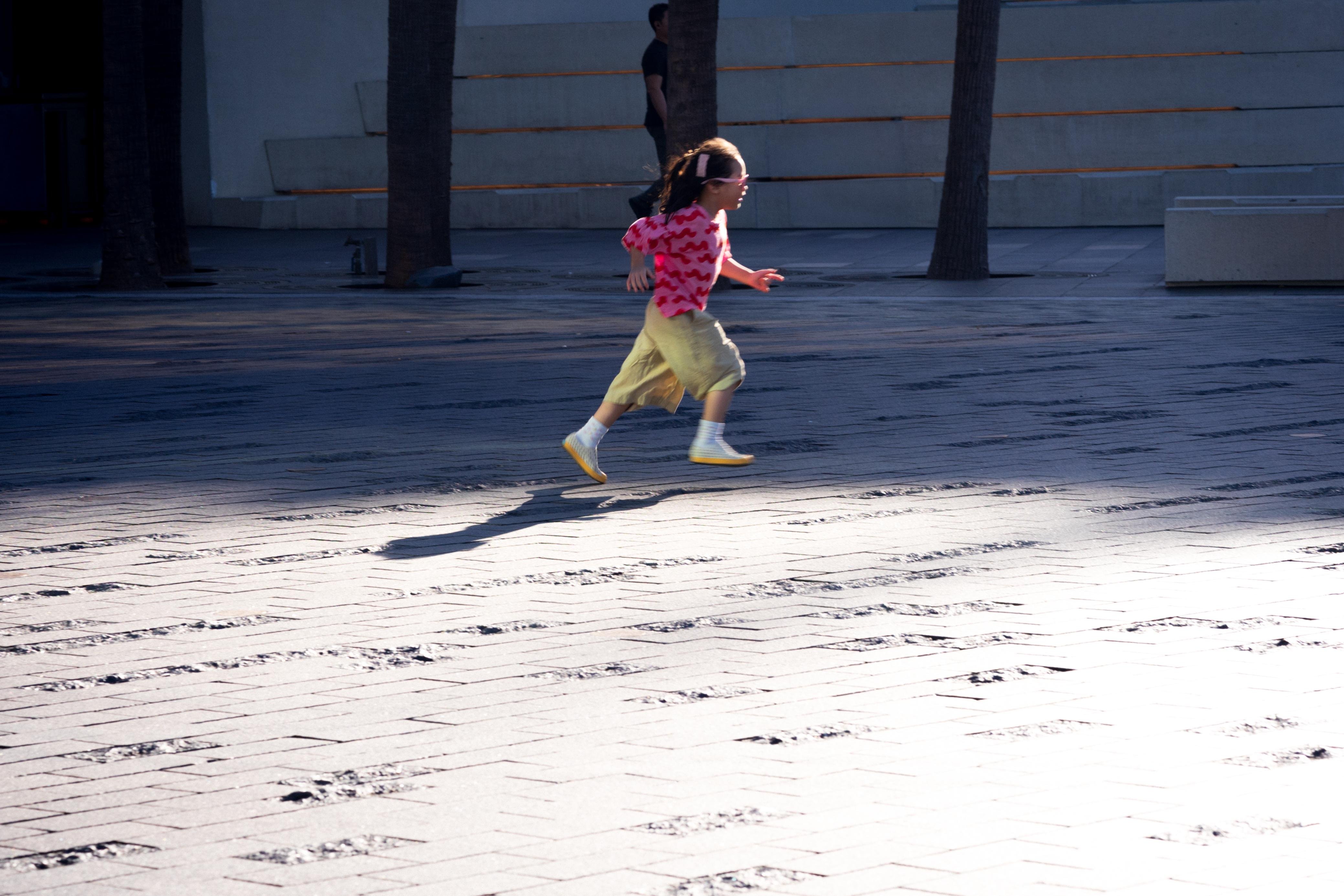 Child with braided hair wearing a pink shirt and light pants running across a sunlit tiled surface. Shadows of trees and other elements fall across the ground. A man stands in the background near concrete steps.