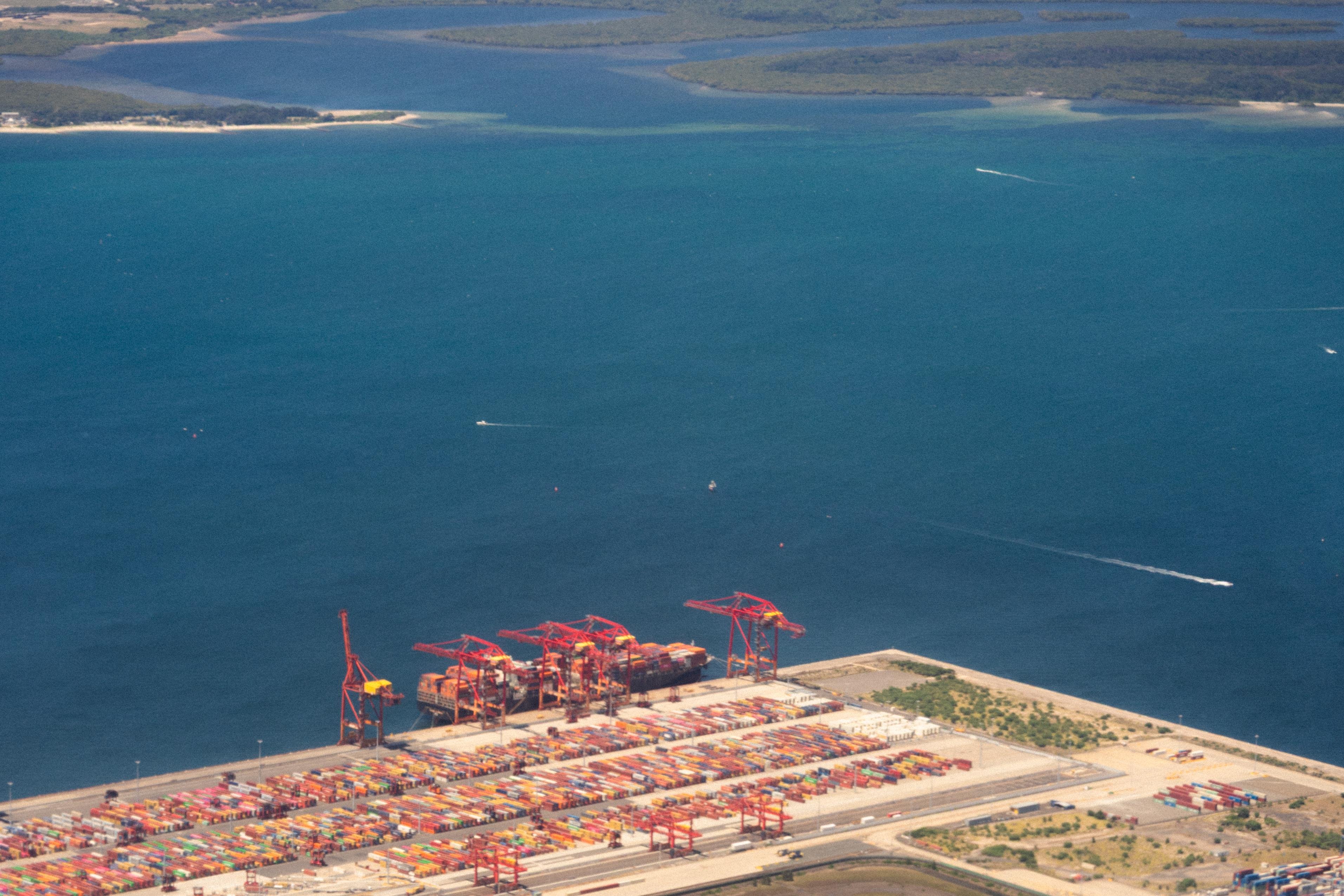 Aerial view of a bustling shipping port featuring numerous colorful shipping containers lined up. Large red cranes are positioned along the dock. The port is adjacent to a vast body of turquoise water with visible land in the background. Small boats leave trails in the water, indicating movement.