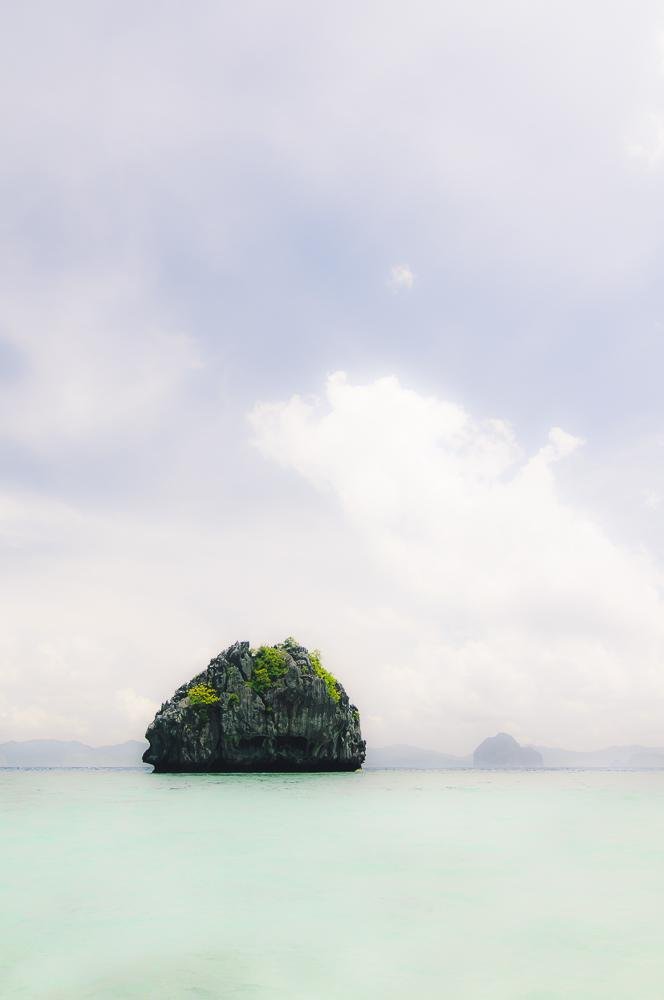 Small rocky islet with patches of green vegetation in calm pale-turquoise water, centered near the horizon, with faint distant islands and a cloudy sky filling most of the frame.