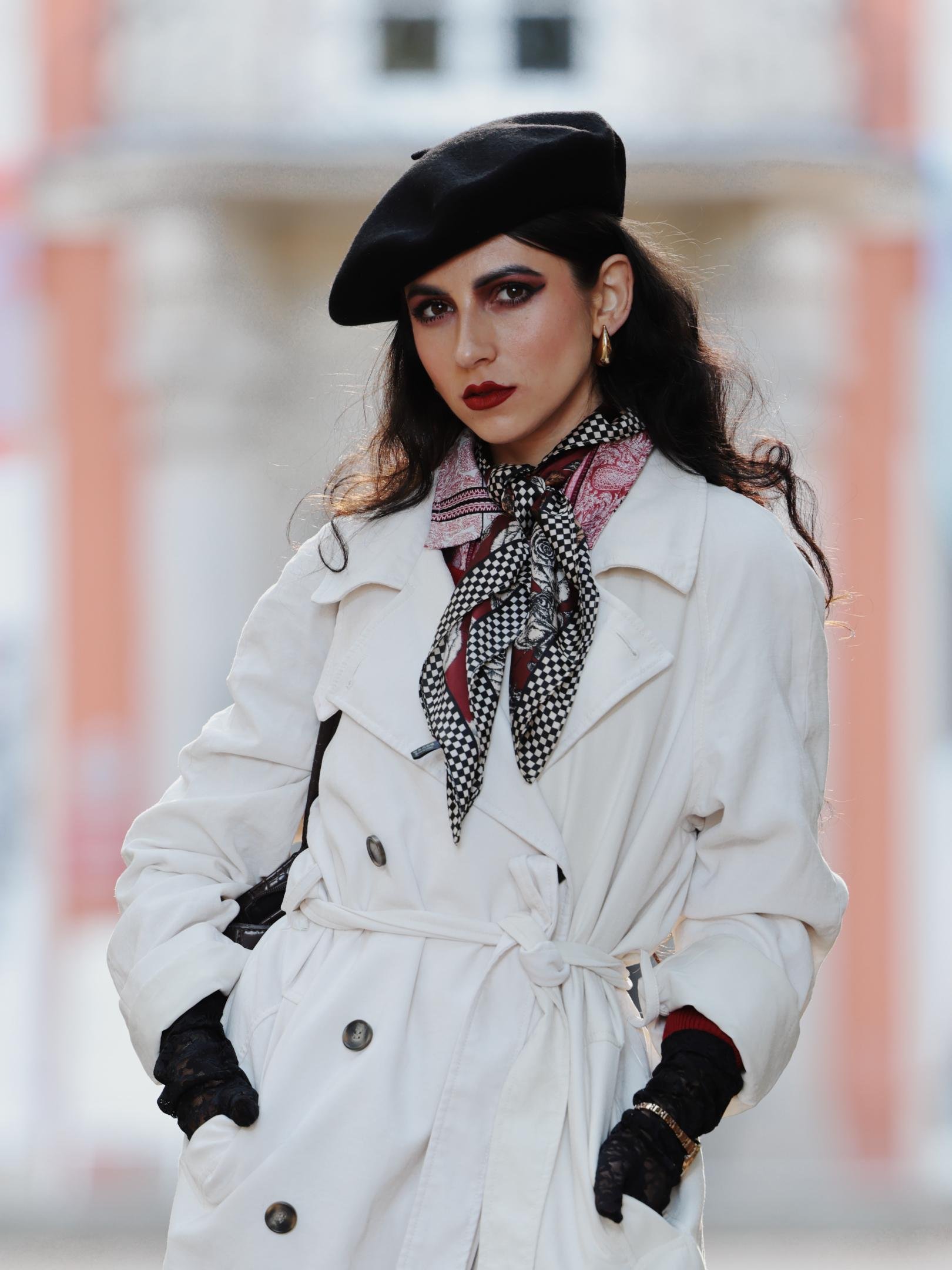 A person wearing a white trench coat, patterned scarf, and black beret stands confidently. They have dark, wavy hair, bold makeup, and are accessorized with earrings and black lace gloves. The background is softly blurred with light and orange tones.