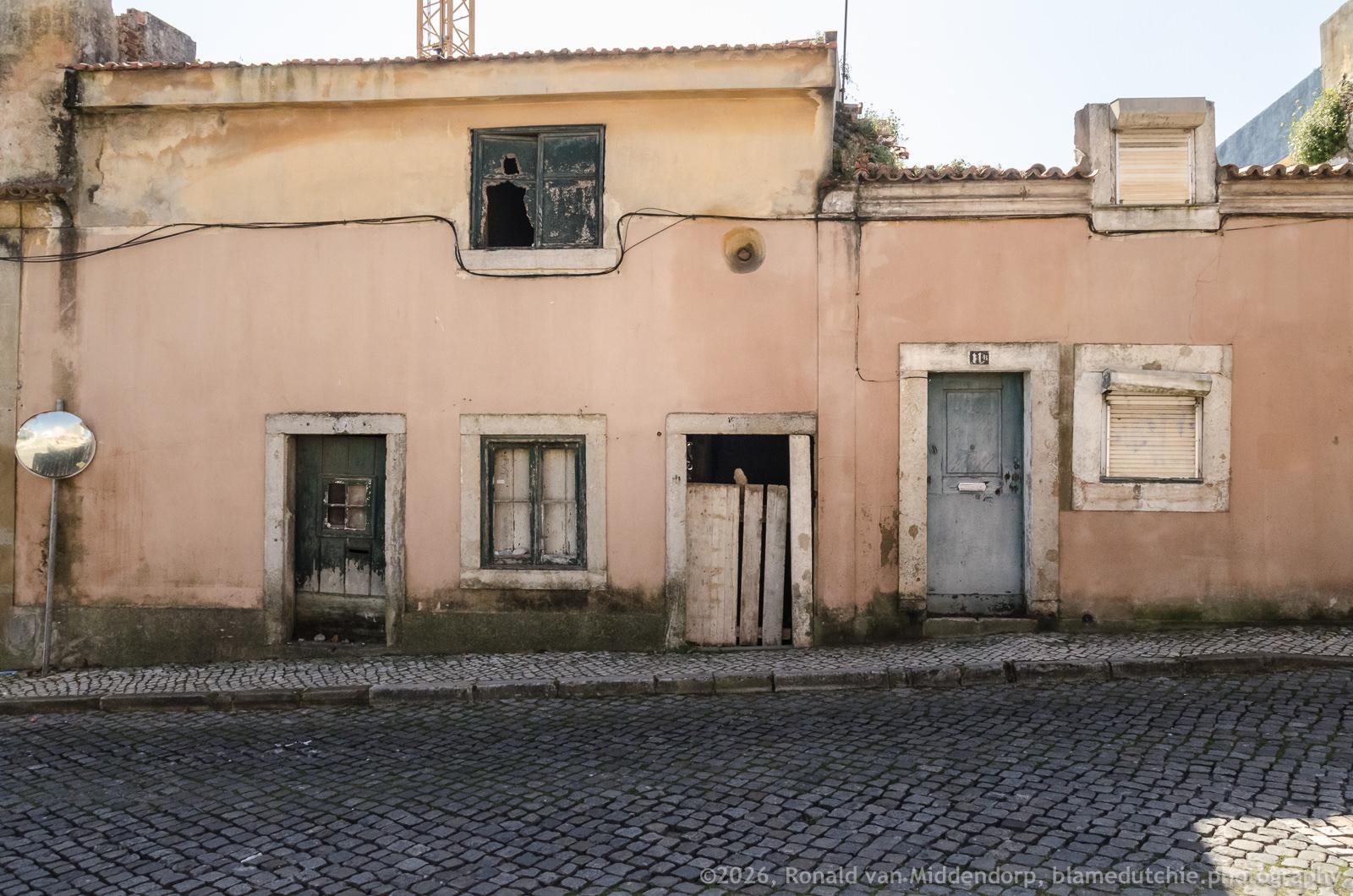 Weathered pink stucco building facade along a cobblestone street, with a broken upper window, two ground-floor windows, a boarded opening, and a gray door; peeling trim, exposed wiring, and a small satellite dish on the left.