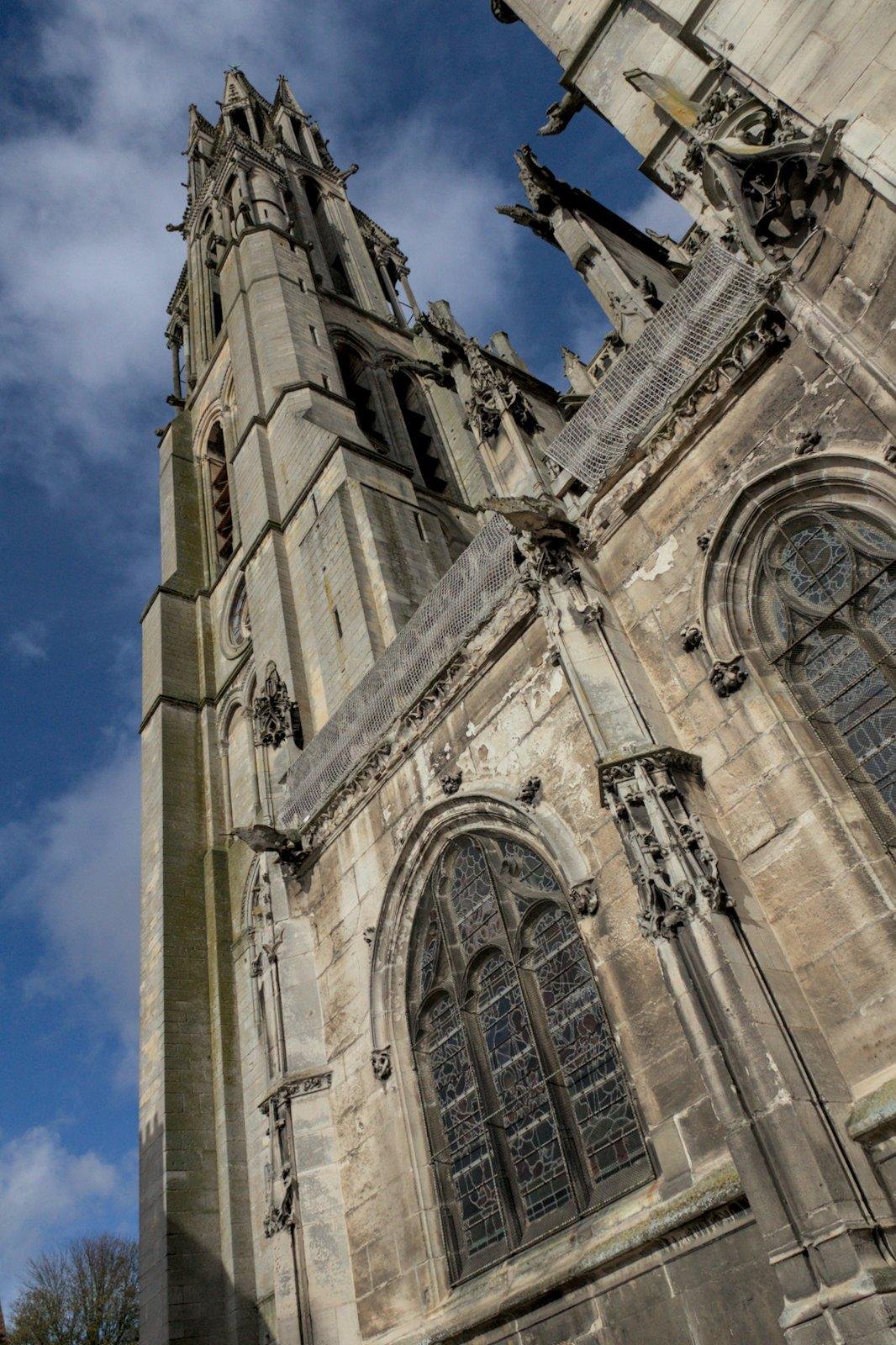 Photo format portrait de La façade de la cathédrale de Senlis en contre-plongée, le ciel est bleu avec quelques nuages, la pierre est beige/jaune, un peu noircie par la crasse