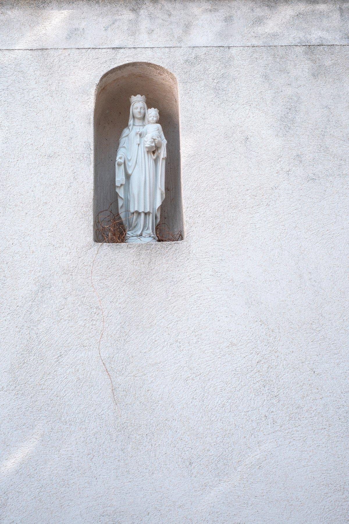 Vertical photo. In a white wall, a small alcove is carved, in which a small white statue of a woman holding a child in her left arm and presenting her right one for us to reach. Probably Mary and Jesus?