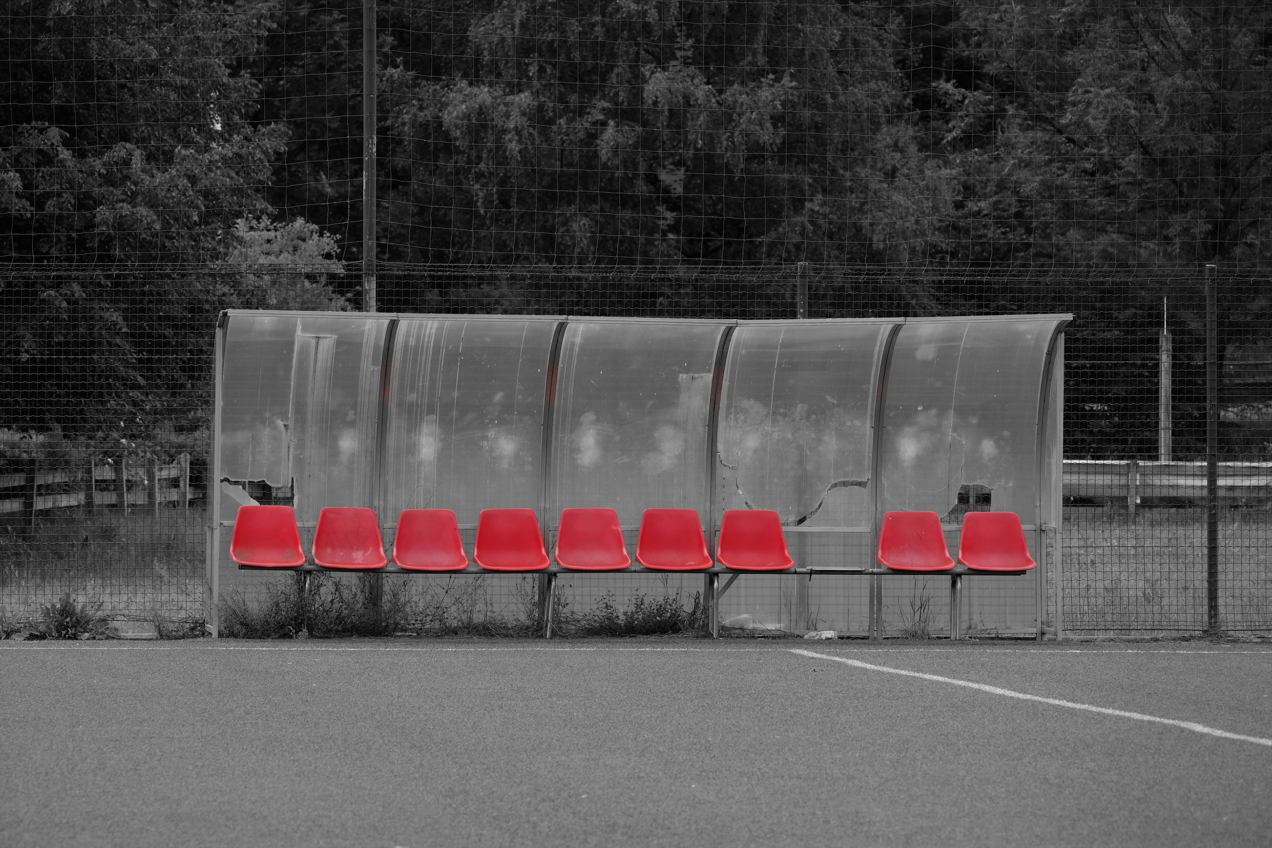 A view from a football field towards the side line where a partially broken plastic roof covers a row of plastic seats, usually used by teams for substitutes und trainers. All seats are empty, one seat is missing from the row.
The complete picture is black and white, except for the seats that show a bright red colour.
