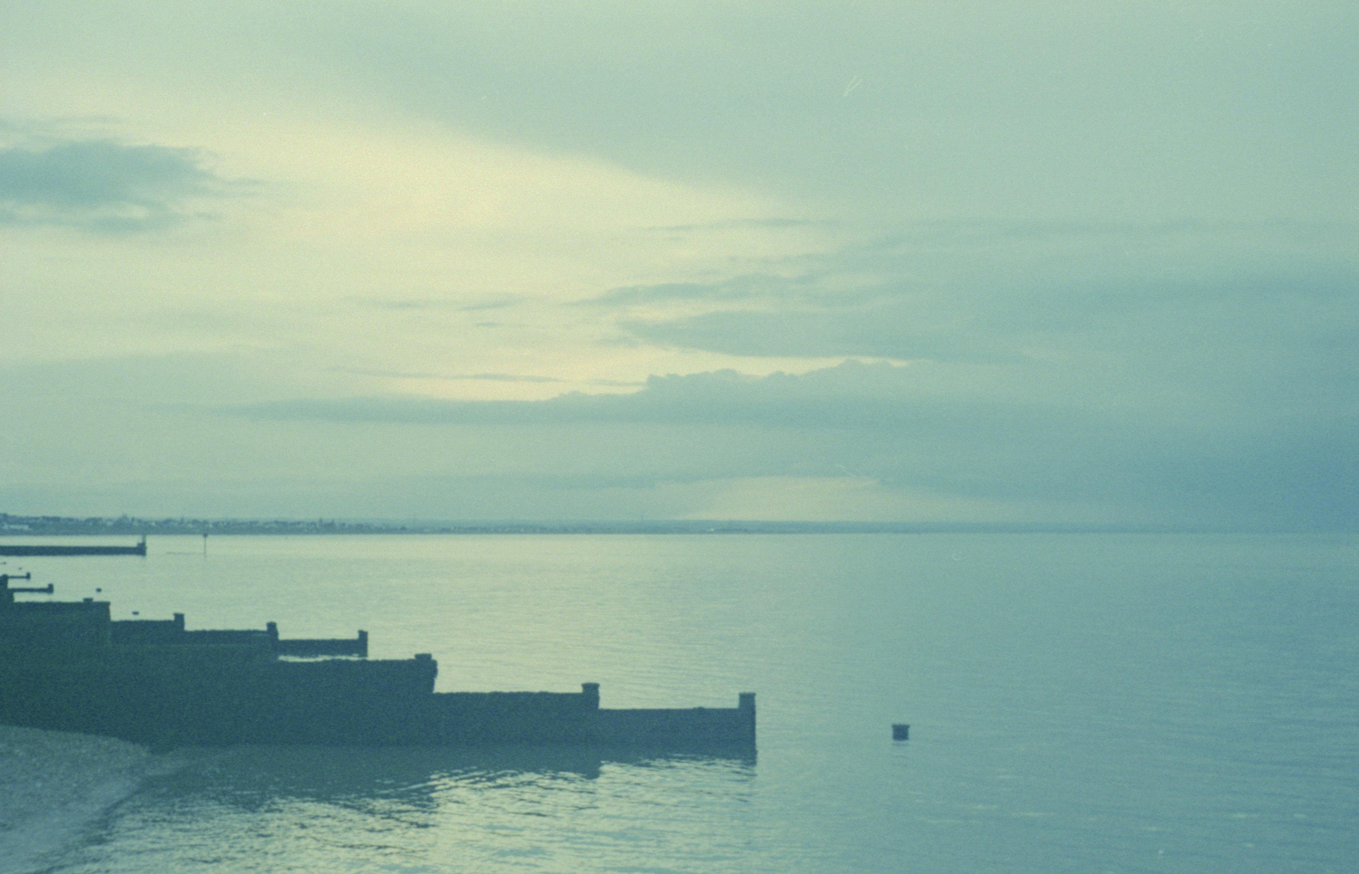 A very simple seascape composition, mostly sea and sky with light, uniform clouds. The left hand shoreline has wooden groynes (sea defences). The whole picture has a blueish tint to it and a slightly faded feel.