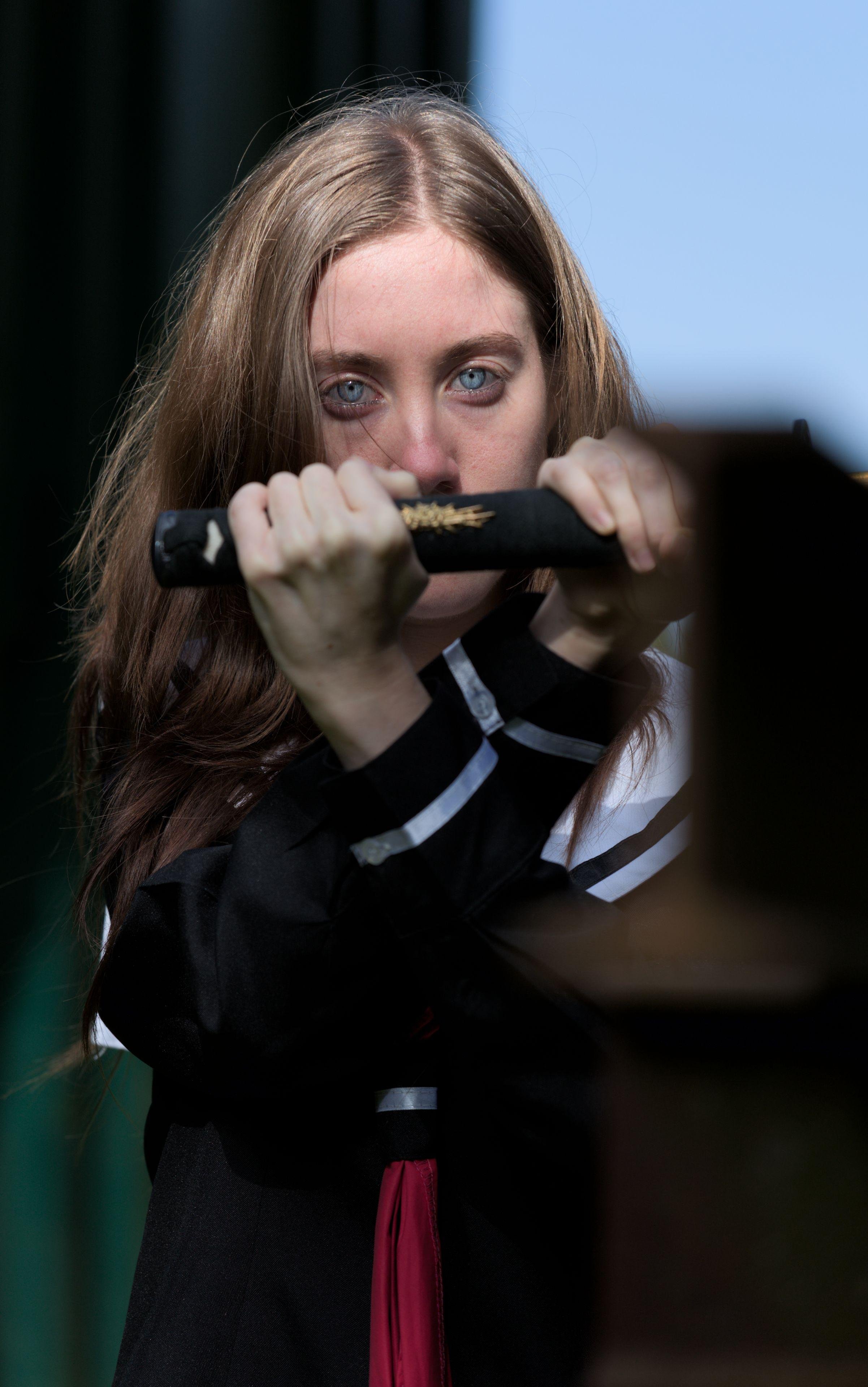 Photo format portrait d’une jeune femme rousse en uniforme d’écolière japonais bleu marine avec un ruban rouge tenant le manche d’un katana en croisant ses bras, les mains de part et d’autre du manche. Le manche cache sa bouche soulignant ses yeux qui regardent droit vers la caméra d’un air menaçant et froid, accentué par la couleur bleu acier de ses yeux