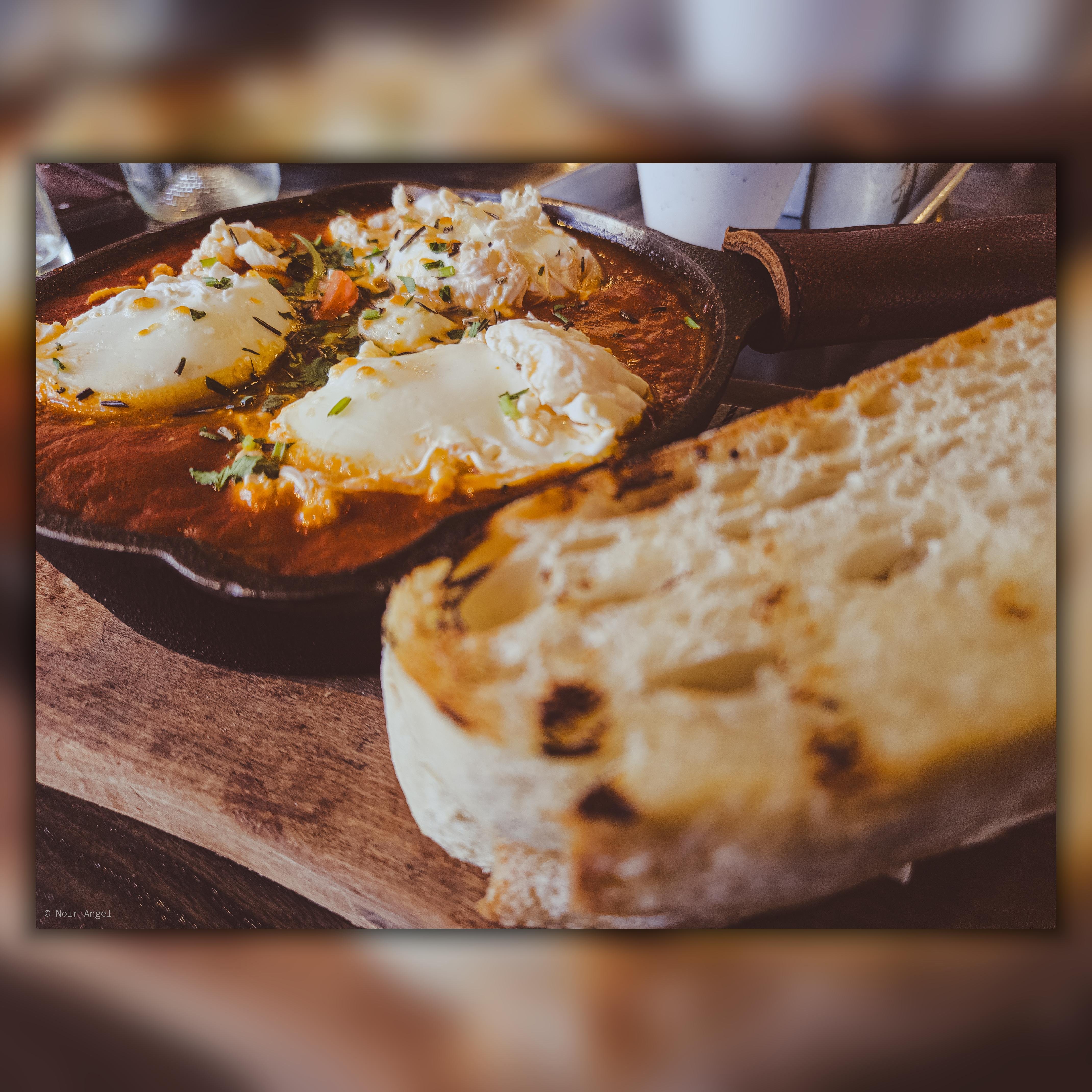 Skillet of shakshuka with poached eggs in a rich tomato sauce, garnished with herbs. Accompanied by a slice of toasted rustic bread on a wooden board. Blurred background with glasses and a white cup.