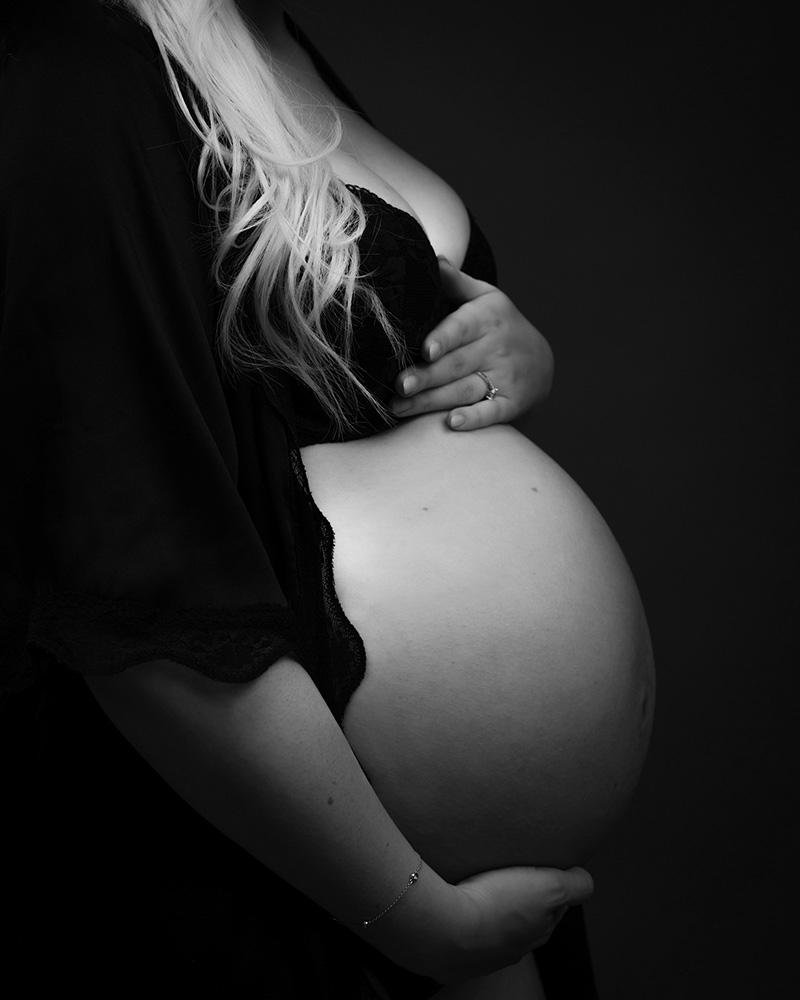 Pregnant woman in a side profile, holding her belly with one hand below and another above. Wearing a dark flowing robe and a bracelet on her right wrist, with long light-colored hair cascading over her shoulder. Black and white image with a dark background.