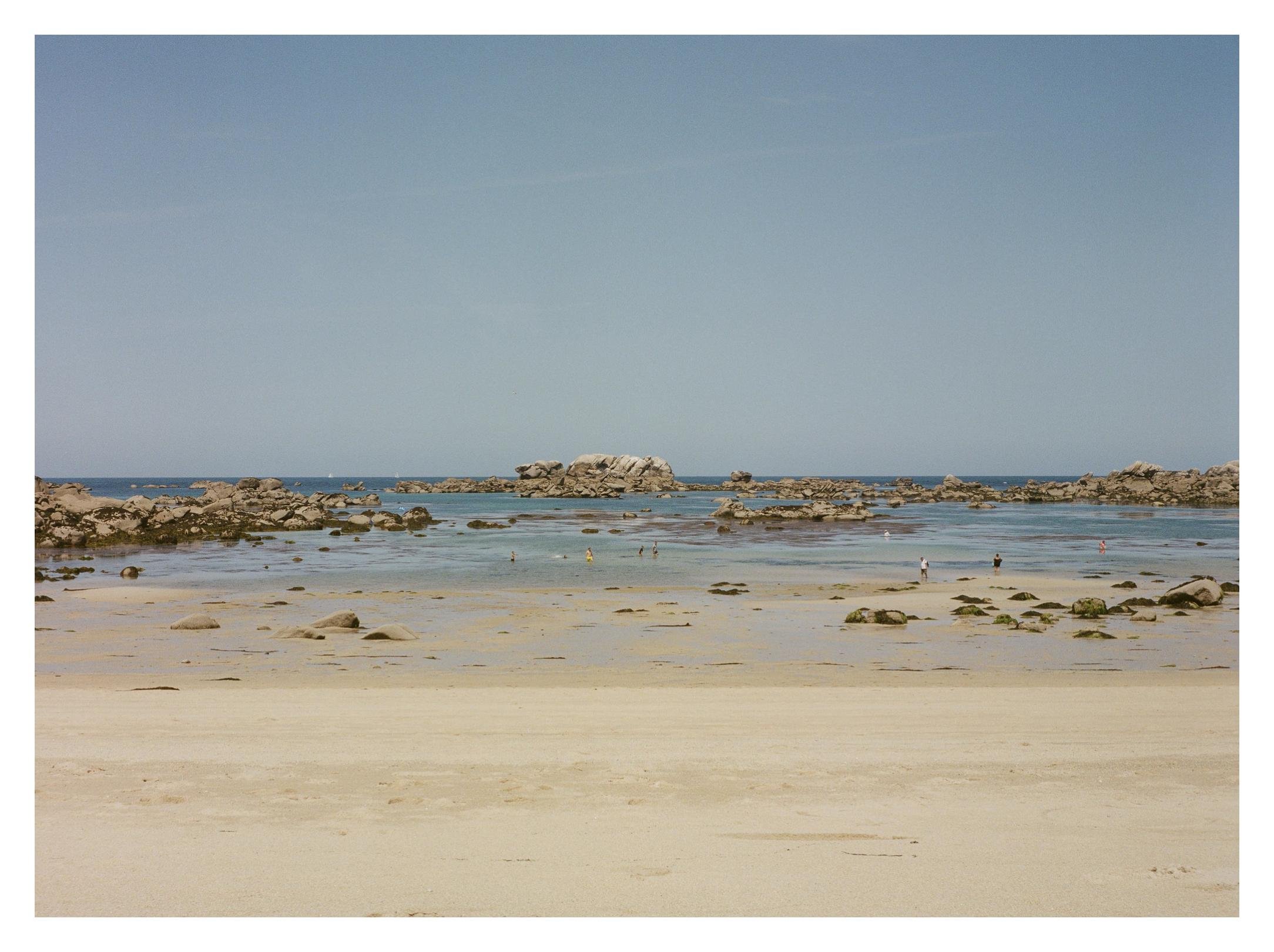 Wide sandy beach in the foreground leading to shallow tidal pools with scattered rocks, a rocky outcrop near the horizon, small people wading in the water, and a clear blue sky above.