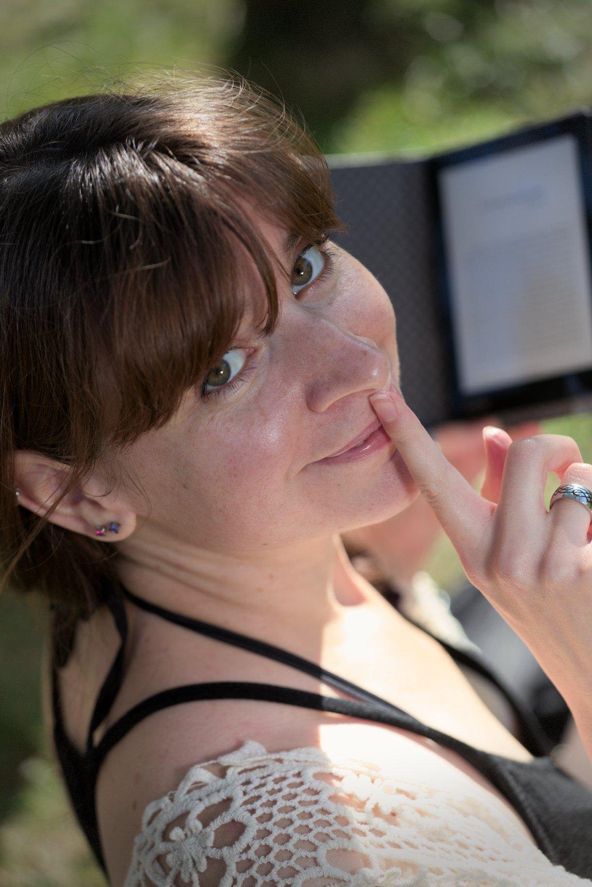 Photo format portrait, en plongée, gros plan du visage d’une jeune femme brune regardant vers la caméra en levant la tête, l’index droit tendu verticalement sur ses lèvres. Elle tiens de la main gauche une liseuse électronique.