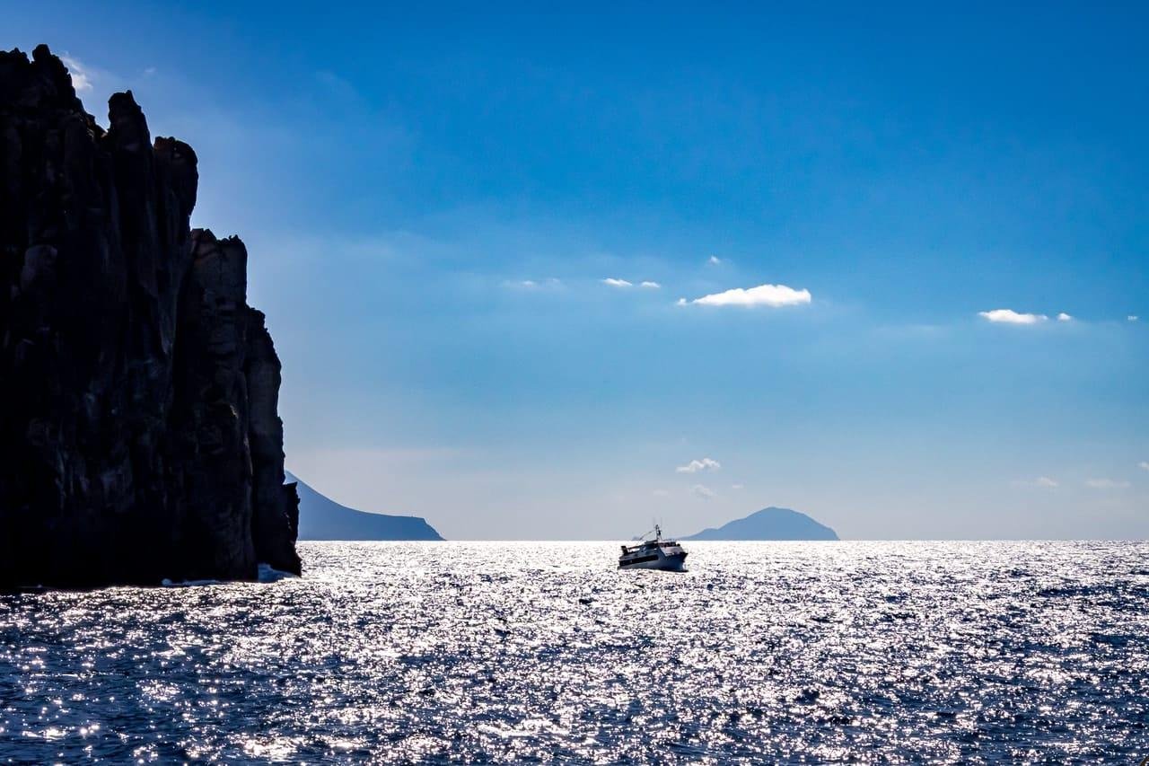 Dark rocky sea cliff on the left, with a small motorboat on sunlit, sparkling water in the center and distant islands on the horizon under a blue sky with a few clouds.